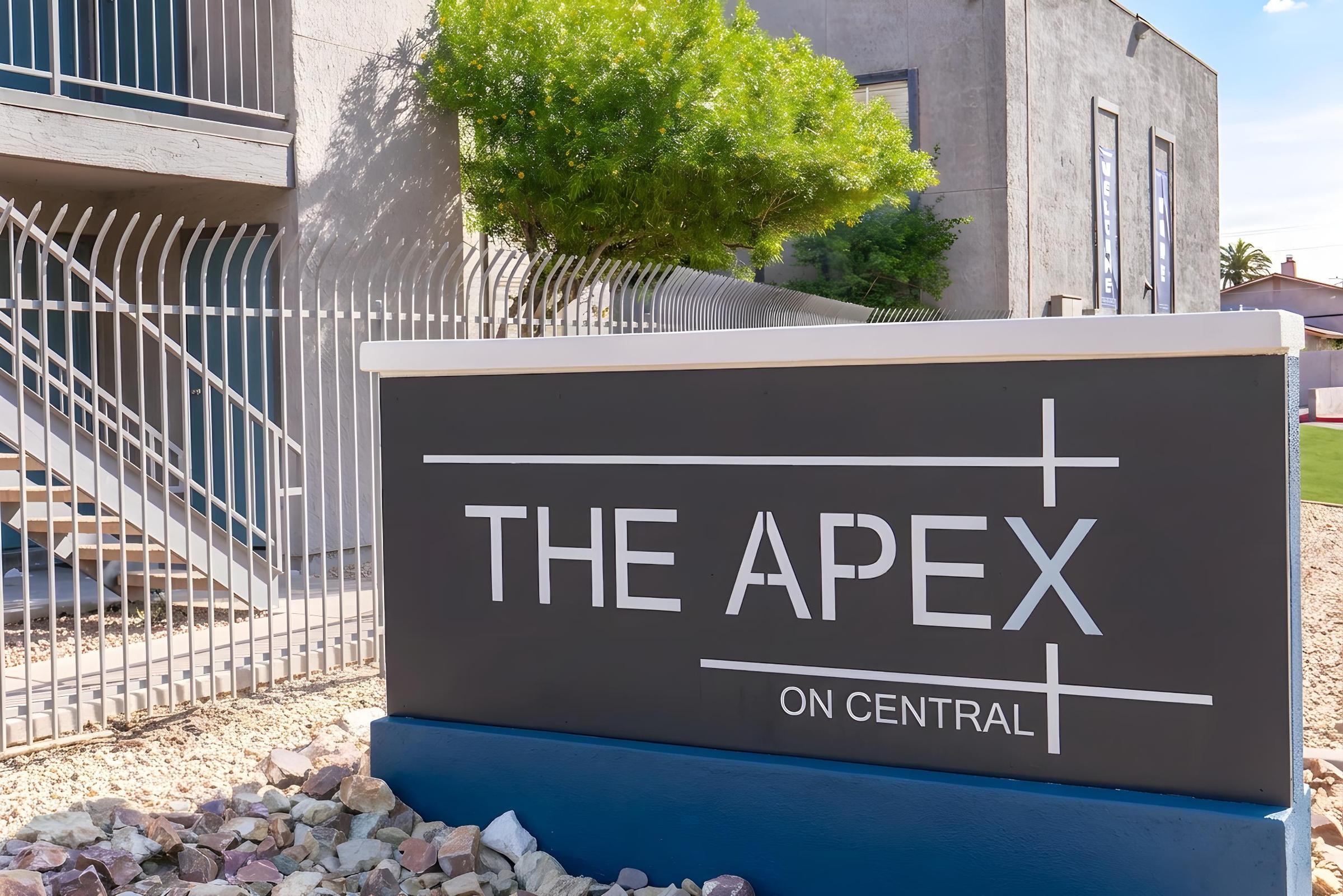 Sign for "The Apex on Central," displayed prominently on a stone base outside a modern building, surrounded by a landscaped area with rocks and greenery. The building features a staircase and railings in the background, indicating an apartment or business complex.