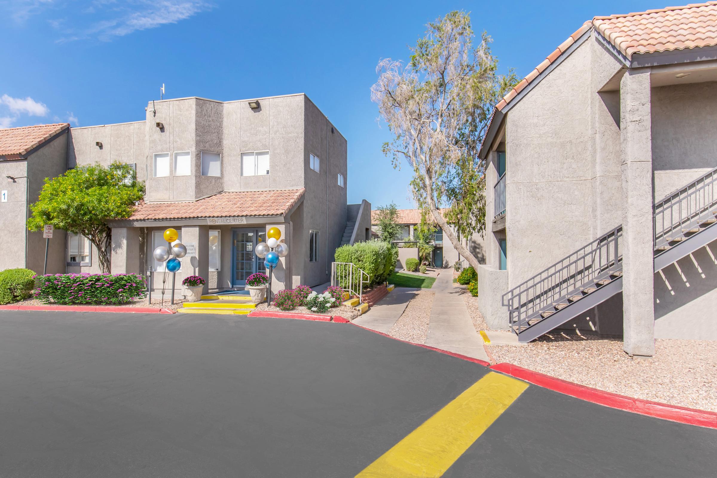 A modern apartment complex with gray buildings, yellow stairs, and colorful balloons at the entrance. Lush greenery and a pathway lead through the complex, with blue skies overhead. This scene conveys a welcoming atmosphere for potential residents.
