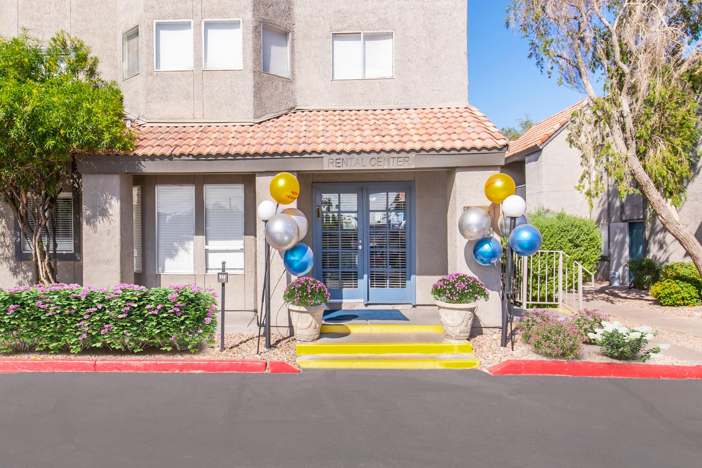 A rental center entrance featuring a tiled roof and large glass doors. Decorative balloons in blue, gold, and silver flank both sides of the door. Flowering plants in pots are placed near the entrance, and the area is well-maintained with a clear blue sky overhead.
