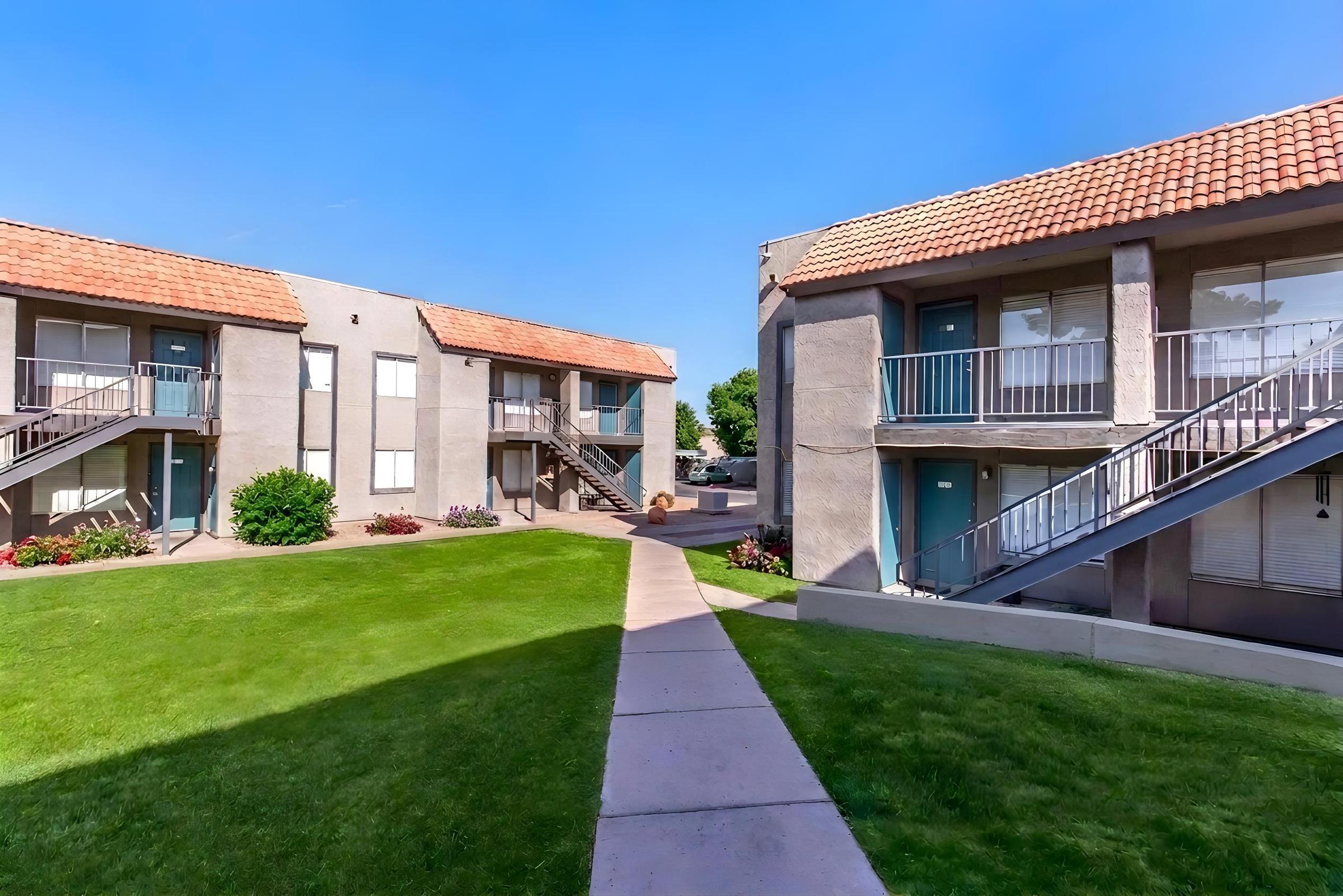 A well-maintained apartment complex with two buildings on either side, featuring a green lawn and a paved walkway in the center. Each building has a set of stairs leading to the upper deck and multiple doors, with colorful flower beds enhancing the landscape under a clear blue sky.