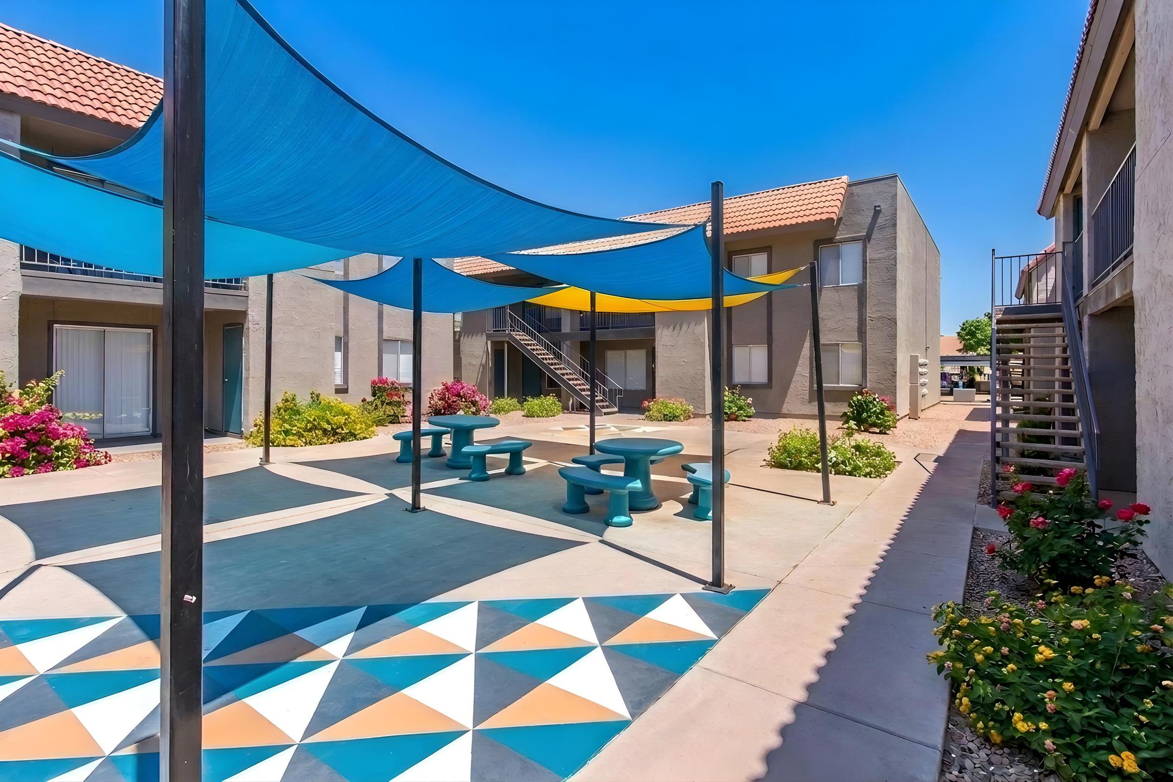 A vibrant outdoor courtyard featuring colorful shade sails, picnic tables, and a decorative tiled floor. Surrounded by blooming shrubs and plants, with apartment buildings visible in the background under a clear blue sky.