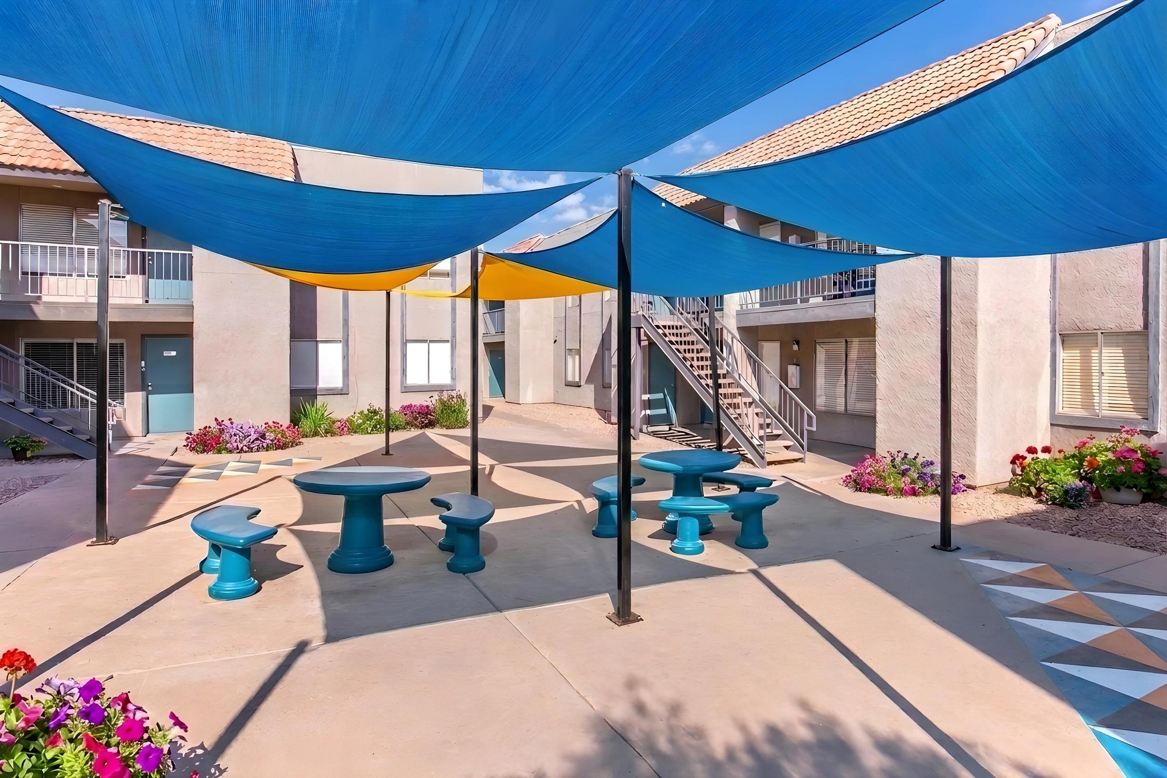 An outdoor courtyard featuring colorful blue and yellow shade sails overhead, with circular tables and stools arranged around a central gathering area. Surrounded by blooming flowers and a light-colored building with balconies, the space offers a vibrant and inviting atmosphere for relaxation or socializing.