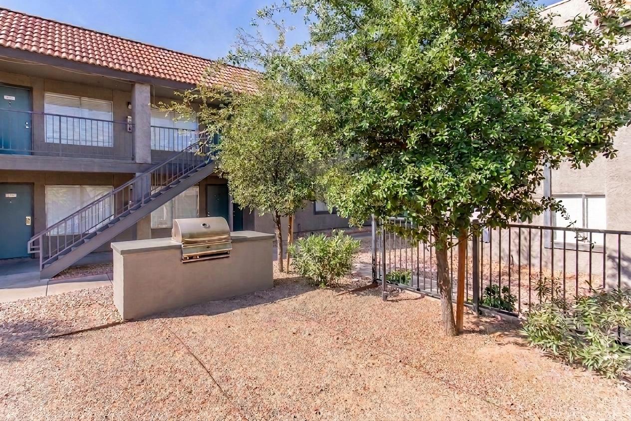 A small outdoor area featuring a barbecue grill, surrounded by gravel and a few plants. In the background are two stories of an apartment building with green doors and a staircase leading to the upper level. There are two trees providing some shade in the area.