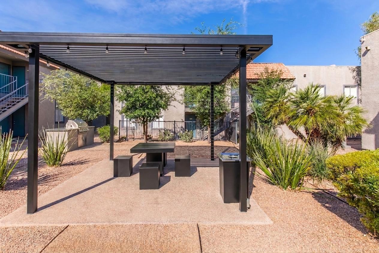 A shaded outdoor area featuring a modern black pergola with a table and chairs, surrounded by landscaped gravel and desert plants. In the background, there's a residential building with stairs and a barbecue grill area visible. Bright blue sky adds a serene ambiance.