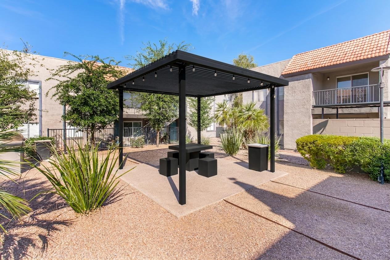 A modern black gazebo with a picnic table and benches, surrounded by landscaped gravel and low-maintenance plants. In the background, there are residential buildings with balconies and green foliage under a clear blue sky. Ideal for outdoor gatherings or relaxation.