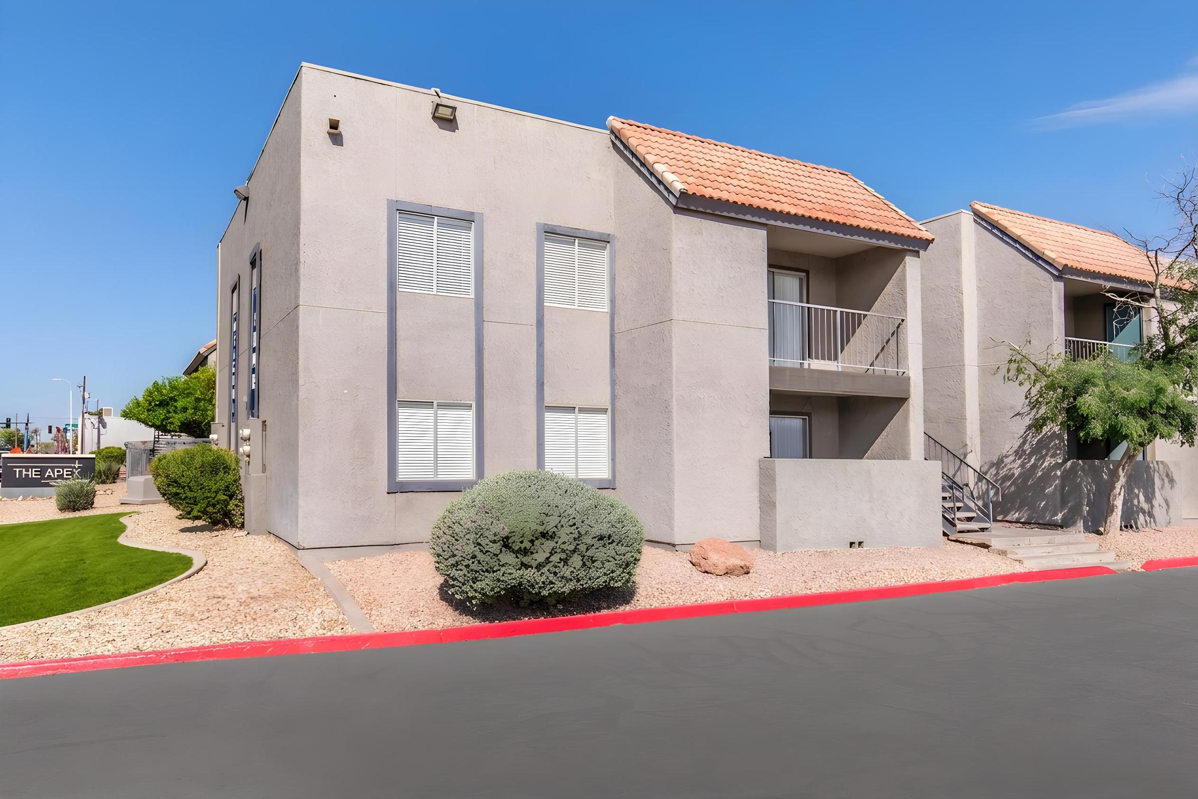 A modern two-story apartment building with a textured gray exterior and red tile roof. The front features white-framed windows and a small balcony. Surrounding landscaping includes gravel and shrubs, with a well-maintained lawn area. A bright blue sky is visible above.