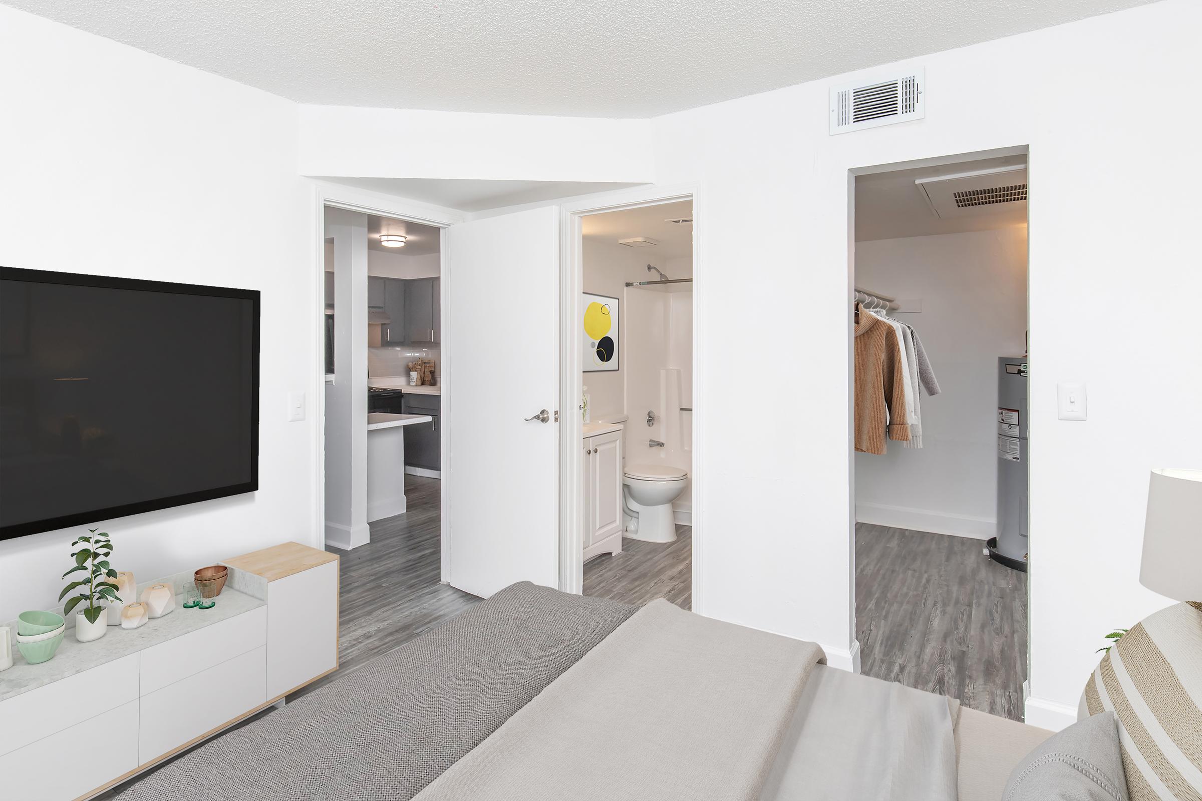 A modern bedroom featuring a bed with gray bedding, a mounted flat-screen TV on the wall, and an open door leading to a bathroom. A closet is visible on the right, and the overall decor is bright and minimalist with light wood flooring and neutral colors.