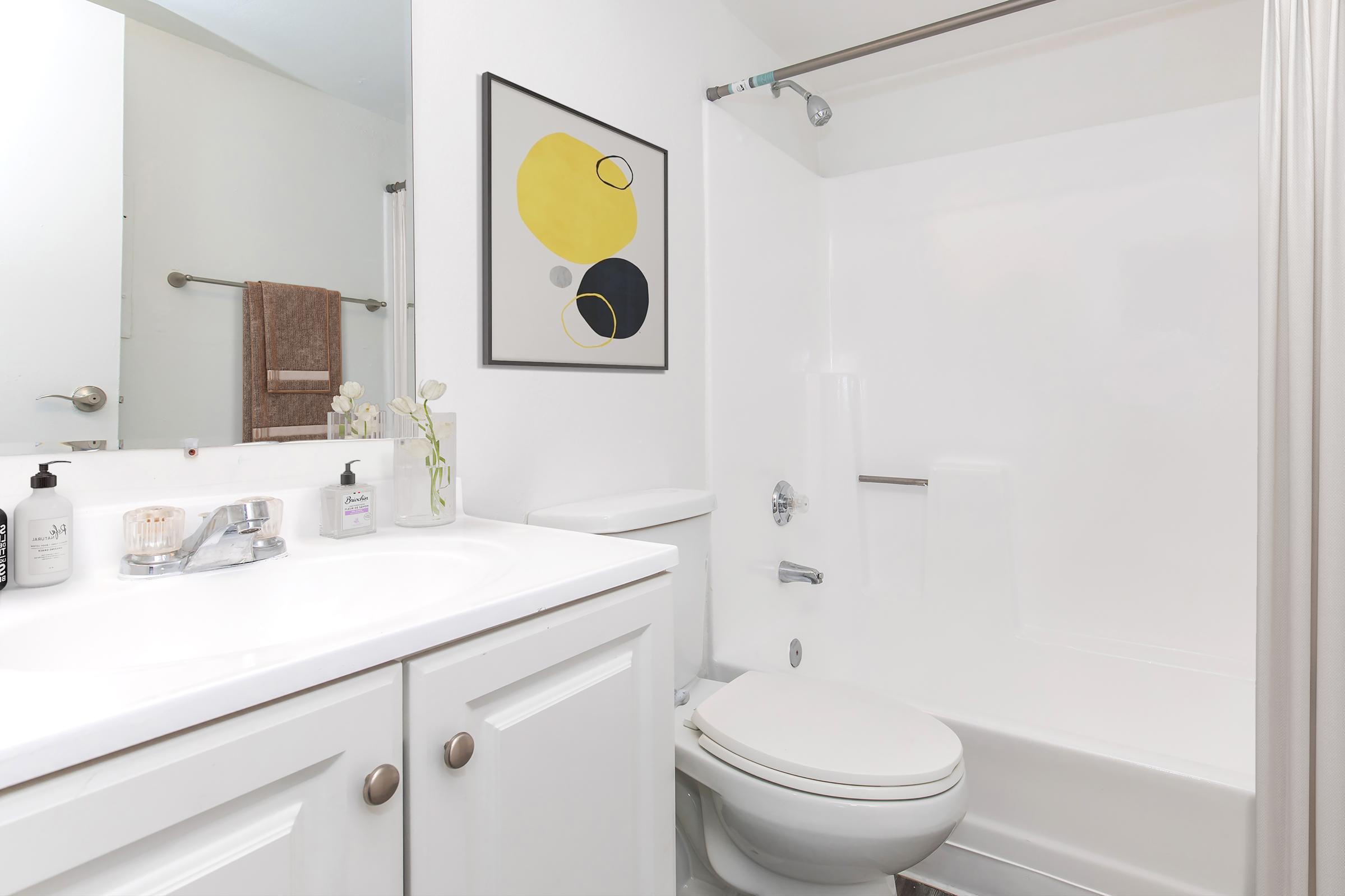 A modern bathroom featuring a white vanity with a silver faucet, a white toilet, and a shower. The walls are painted white, and there's a piece of abstract art with yellow and black shapes hanging on the wall. A towel rack holds a brown towel, and a small vase with flowers adds a decorative touch.
