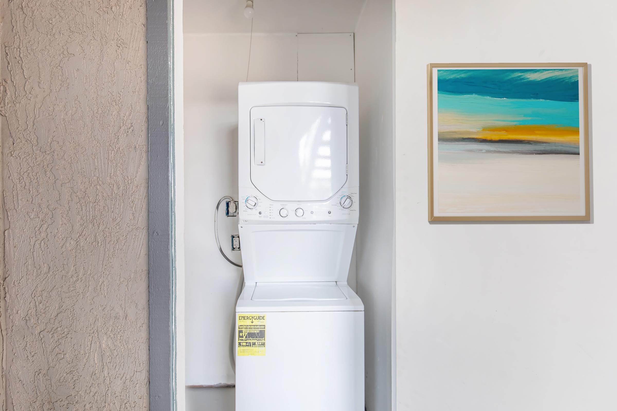 A compact laundry area featuring a stacked washer and dryer unit with a warning label. Adjacent to it is a minimalist framed artwork displaying abstract colors of blue, green, and tan. The walls are in neutral tones, creating a simple and functional space.