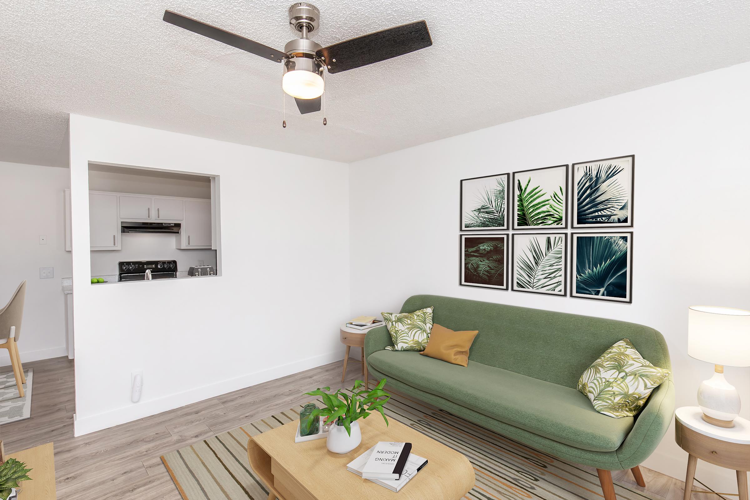 A bright, modern living room featuring a green sofa with decorative pillows, a wooden coffee table, and a small indoor plant. Wall art depicting tropical leaves hangs above the sofa. A ceiling fan adds a stylish touch, and a glimpse of the kitchen is visible through an open space in the wall.