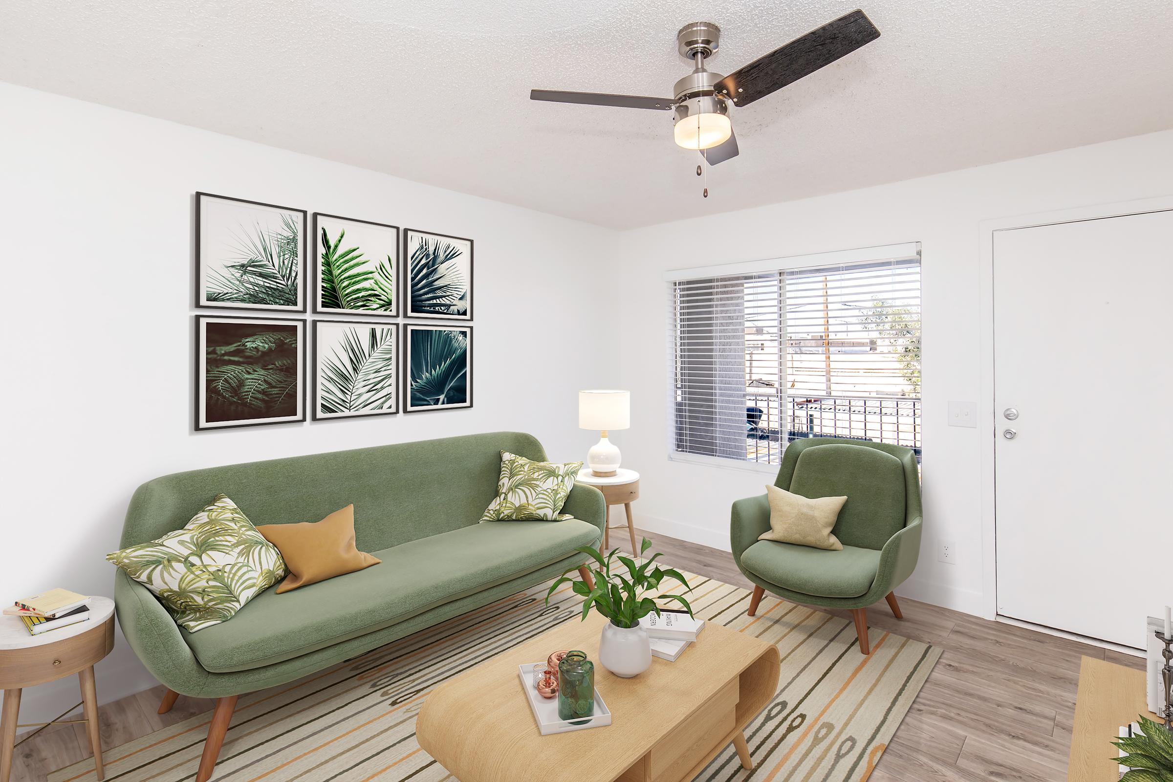 A modern living room featuring a green sofa with cushions, a light wood coffee table, and a small side table with a lamp. The walls are adorned with framed palm leaf art, and a ceiling fan hangs above. A bright window offers natural light, and a small potted plant sits on the table.