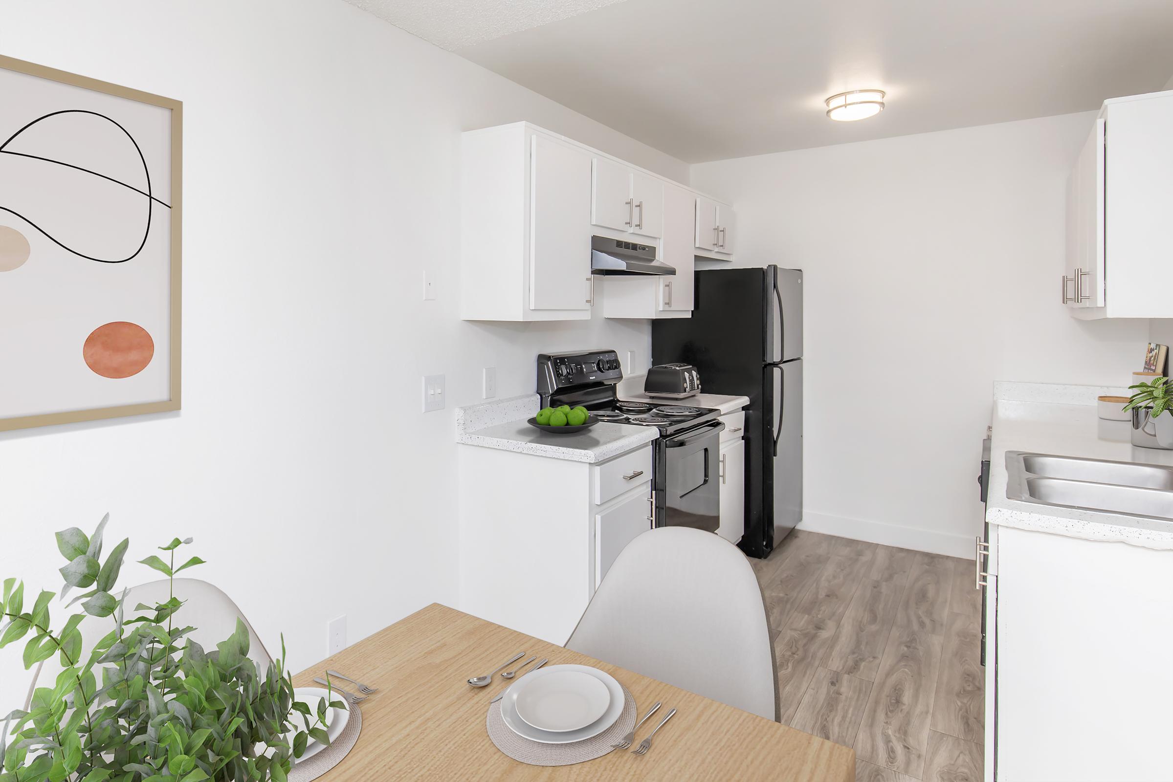 A modern kitchen with white cabinets, black appliances, and a light countertop. A dining table with two plates and a plant sits in the foreground. An abstract art piece hangs on the wall, adding a contemporary touch to the space. The overall atmosphere is bright and inviting.