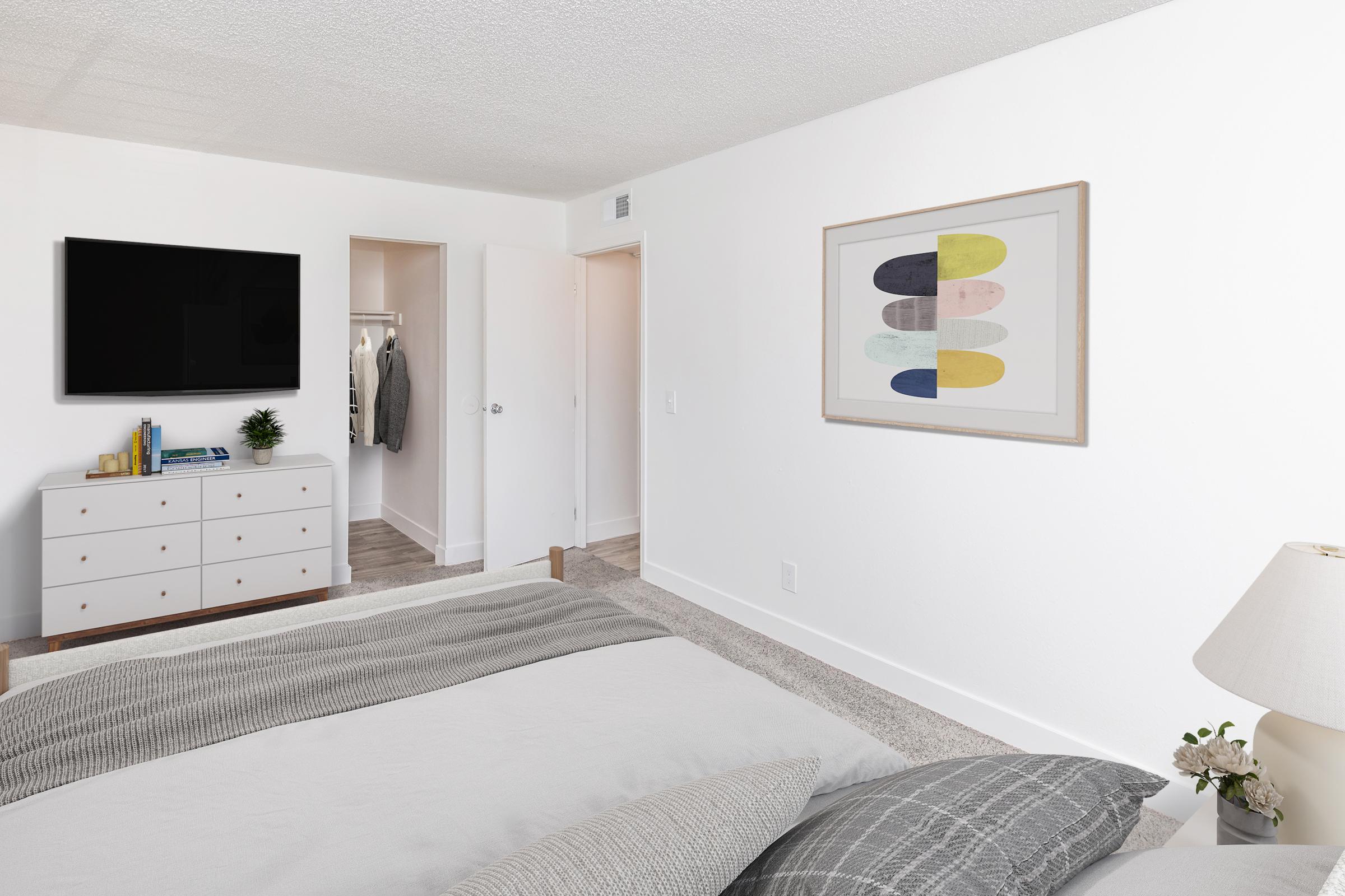 A modern, minimalist bedroom featuring a bed with gray bedding, a white dresser with decorative items, a mounted flat-screen TV, and a framed artwork on the wall. A closet door is partially open, revealing clothes inside. Soft lighting and neutral colors create a calm atmosphere.