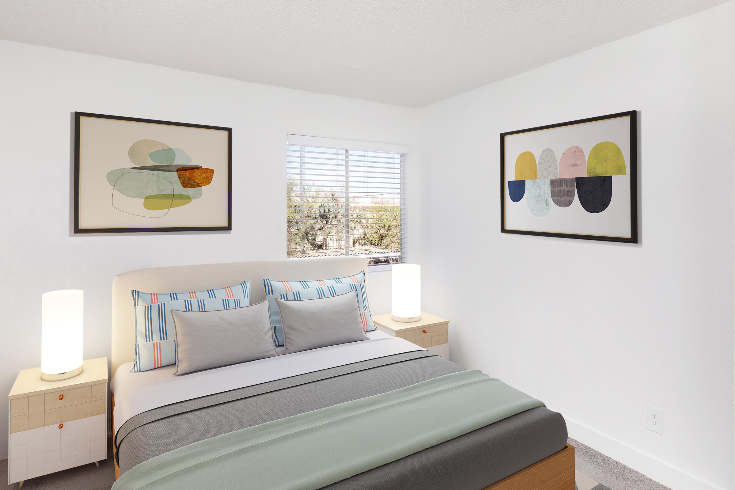 A modern bedroom featuring a bed with light gray and mint green bedding, two decorative pillows, and bedside tables with table lamps. The walls are white, adorned with abstract art pieces in neutral colors. A window with blinds provides natural light, offering a view of a desert landscape outside.