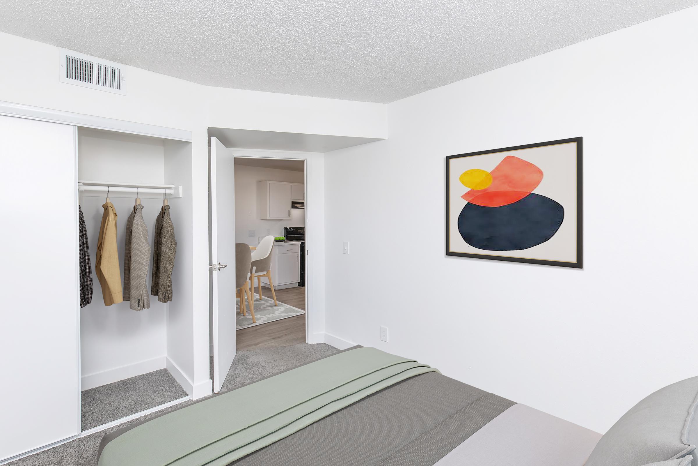 A modern bedroom featuring a neatly made bed with green and gray bedding. A minimalist abstract painting hangs on the white wall. An open closet reveals hanging clothes, and a doorway leads to a kitchen area in the background. Natural light brightens the space, creating a clean and inviting atmosphere.