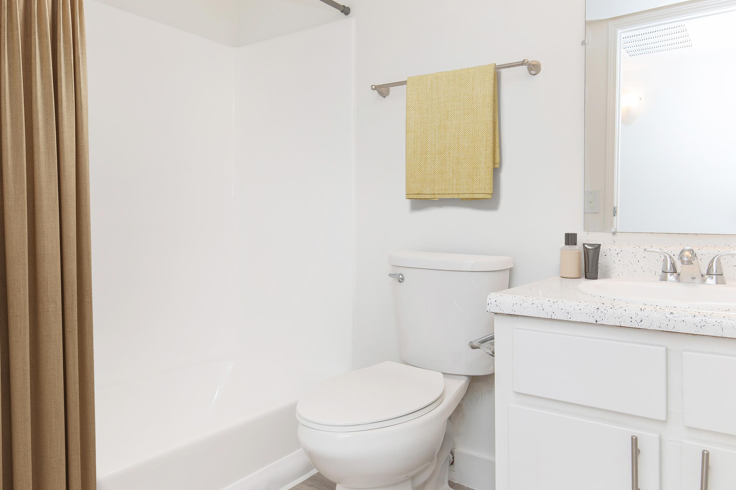 A clean, modern bathroom featuring a bathtub with a shower curtain, a white toilet, and a single sink vanity with a speckled countertop. A yellow towel hangs on a rod, and there's a mirror above the sink. The walls are painted white, creating a bright and fresh atmosphere.
