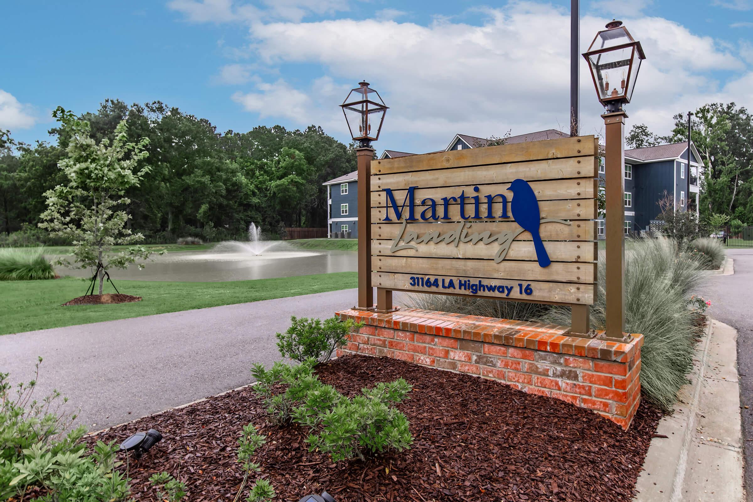Sign for Martin Landing located at 31164 LA Highway 16, featuring a wooden design with blue lettering and a silhouette of a bird. The background includes a landscaped area with greenery and a fountain.