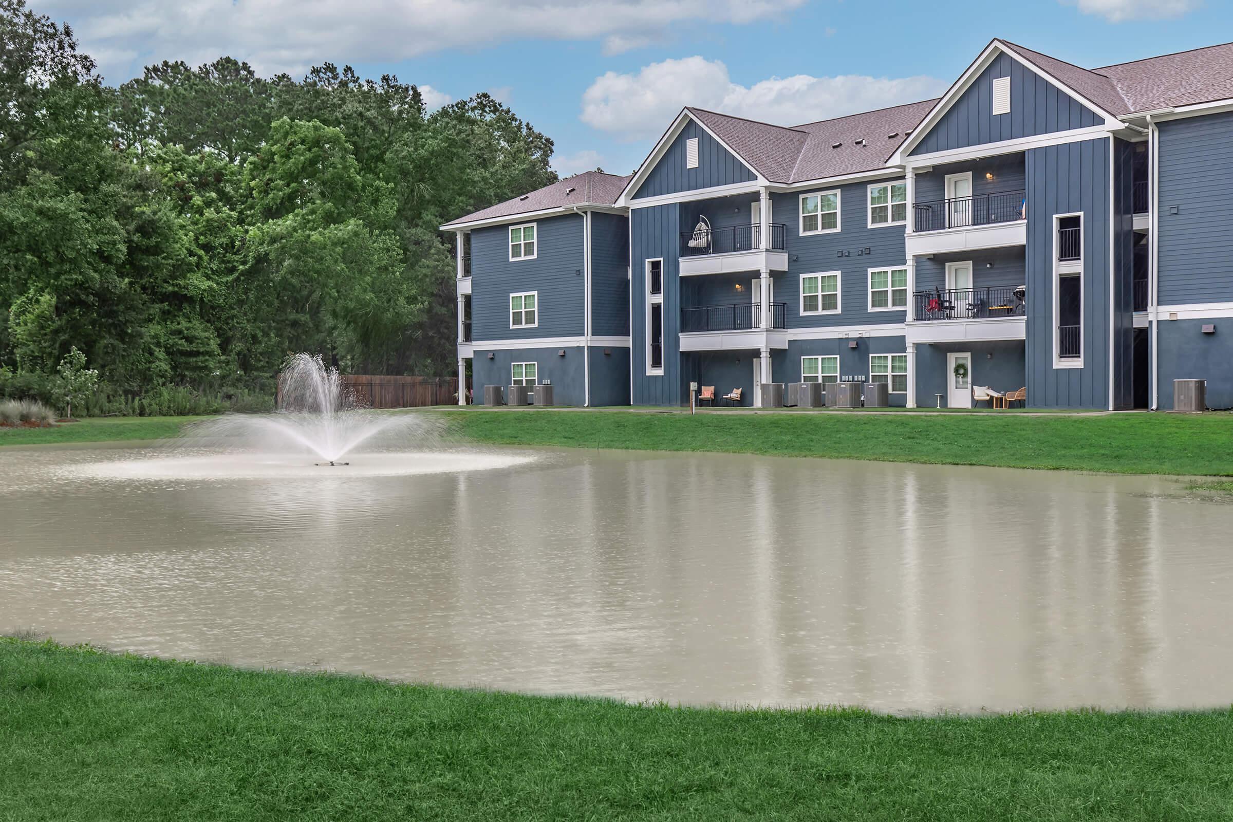 A peaceful residential scene featuring an apartment building with blue siding alongside a pond. The pond has a fountain creating a gentle spray of water, surrounded by green grass and trees in the background, under a partly cloudy sky. The setting conveys a serene living environment.