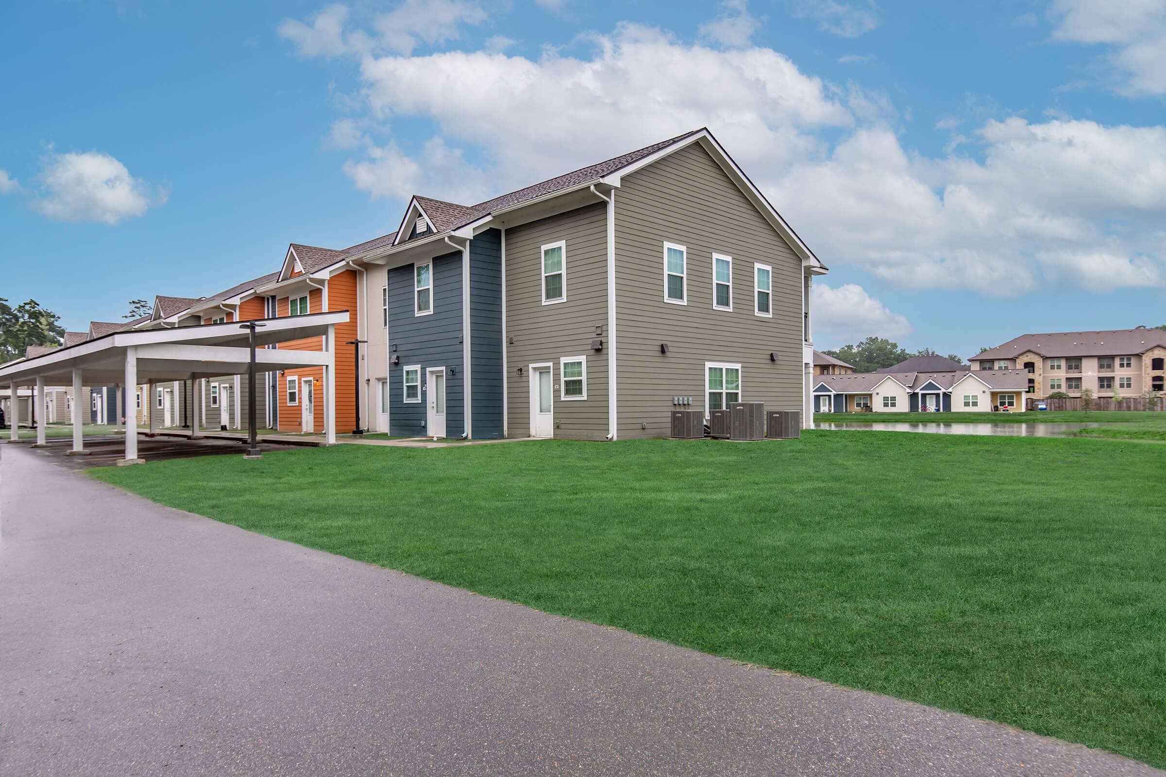 A modern residential building featuring multiple units with varied exterior colors, including orange and blue, set on a well-maintained green lawn. The view is from a paved walkway leading to the building, with overcast skies in the background and more apartments visible in the distance.