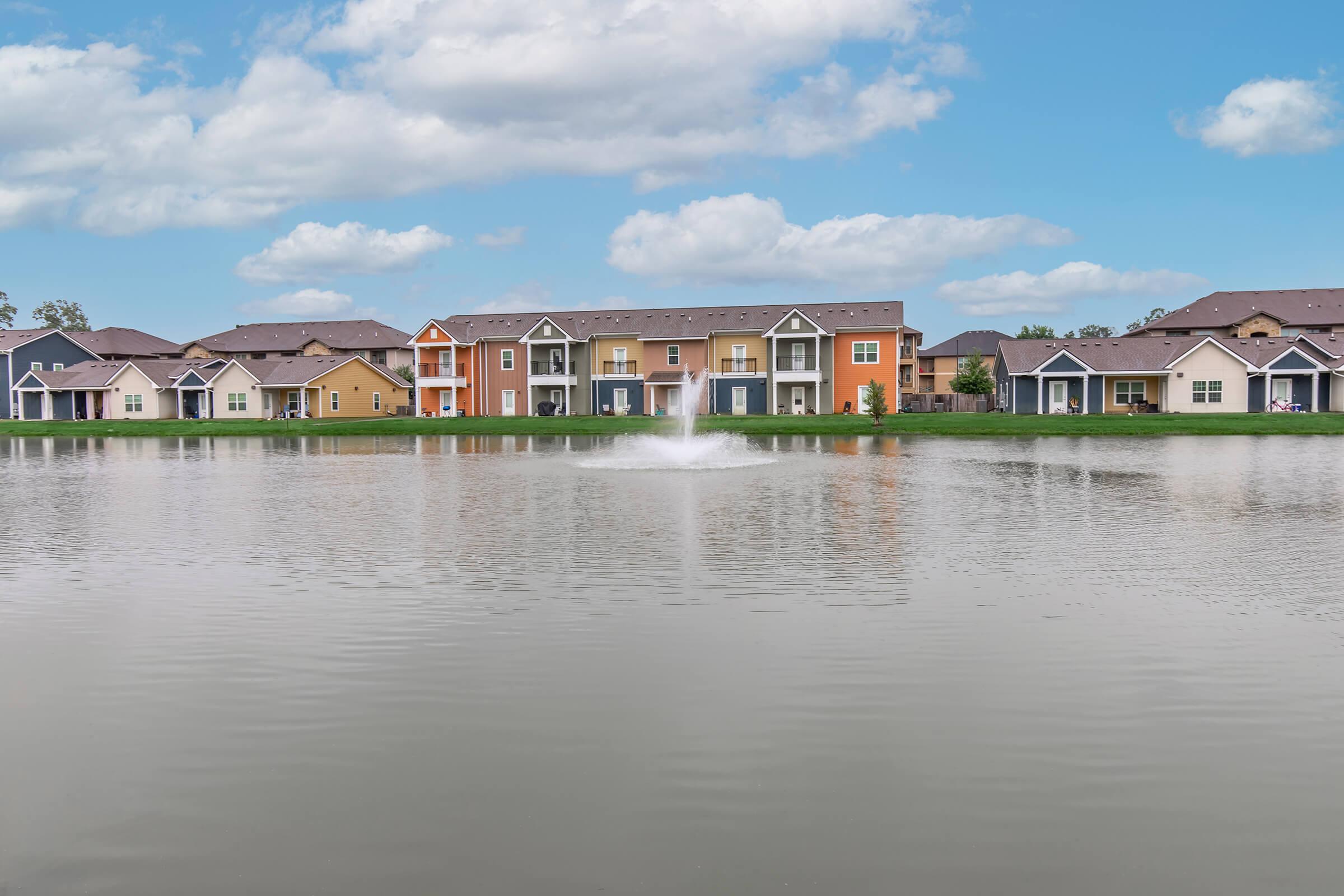 A serene lake surrounded by colorful residential buildings. In the center, a fountain sprays water into the air, creating a peaceful atmosphere. The sky above is partly cloudy, enhancing the tranquil setting of the community.