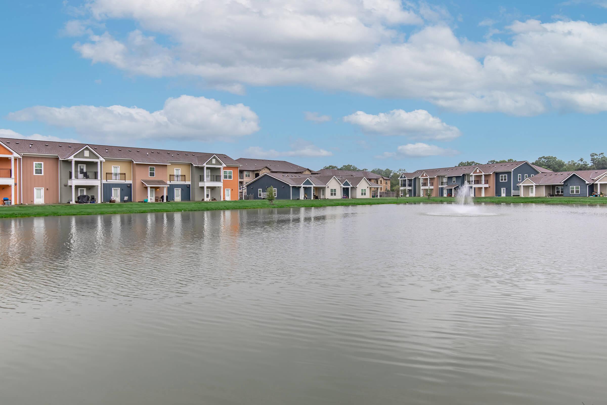 A serene view of a residential community with colorful apartment buildings lining the shore of a calm pond. The water reflects the sky and features a small fountain, surrounded by green grass and trees, under a partly cloudy sky.