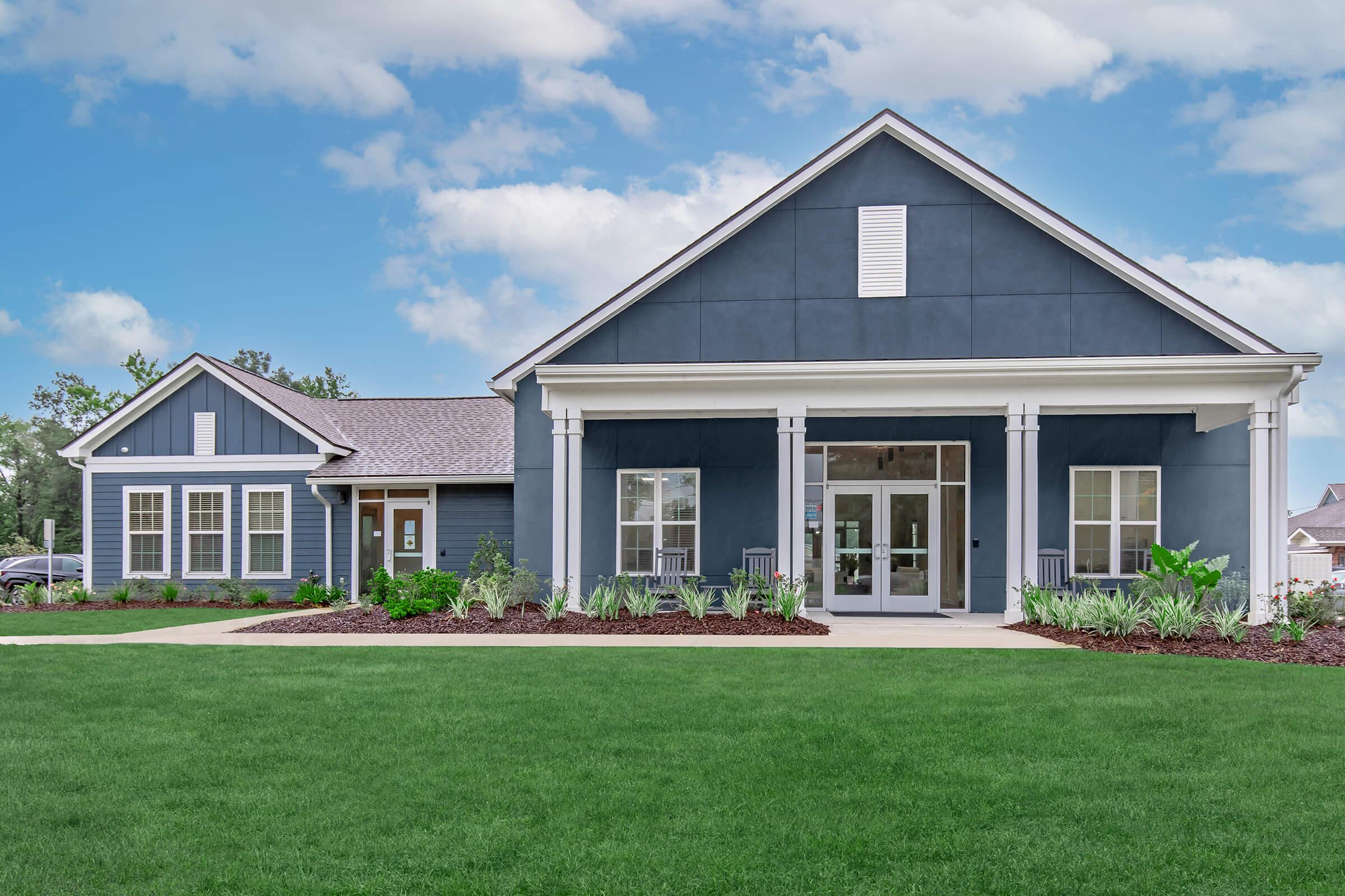 A modern two-story house with a blue exterior, featuring a welcoming front porch and large windows. The landscaped yard is well-maintained with green grass and decorative plants, set against a bright sky with scattered clouds.