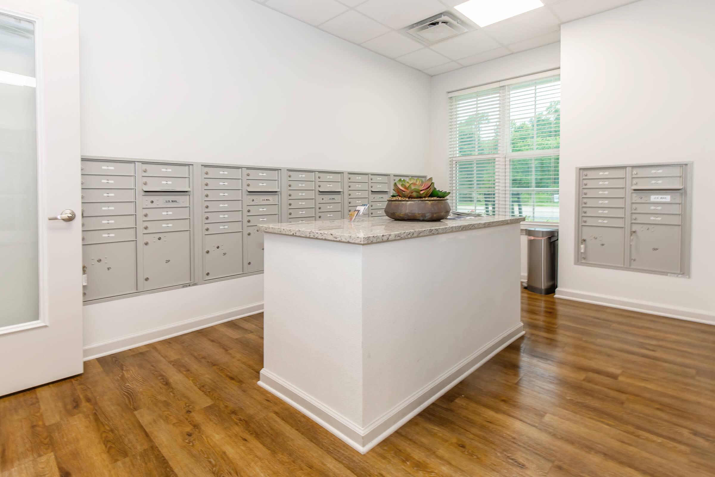 Interior of a mailbox area featuring a central counter with a decorative plant. Surrounding walls have multiple gray mailboxes. There is a large window providing natural light, and the floor is a warm wood finish. The space appears clean and organized, suitable for a residential or office building.