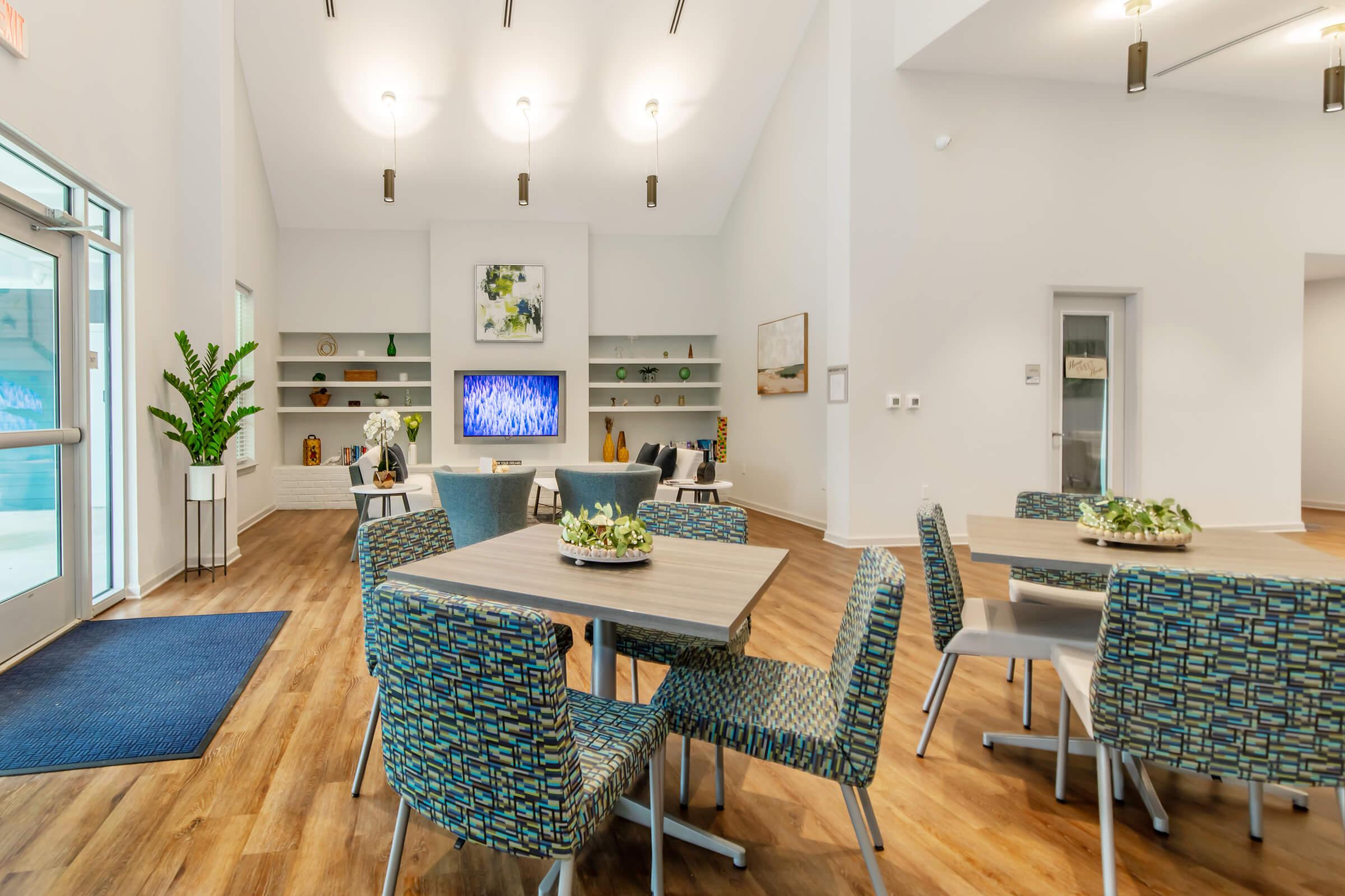 Modern interior of a common area featuring light-colored walls, wooden flooring, and elegant lighting. There are several tables with patterned chairs arranged around, a large TV mounted on the wall displaying blue visuals, and decorative plants enhancing the space. A welcoming atmosphere with minimalistic decor.