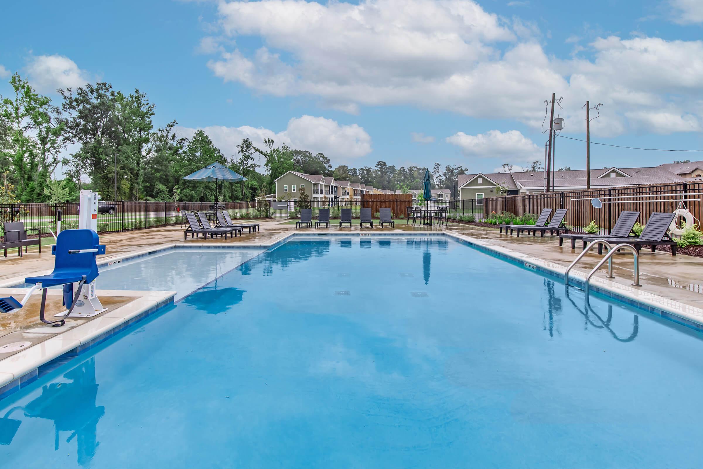 A clear blue swimming pool surrounded by lounge chairs and umbrellas. Green trees in the background, with buildings visible beyond the pool area under a partly cloudy sky.