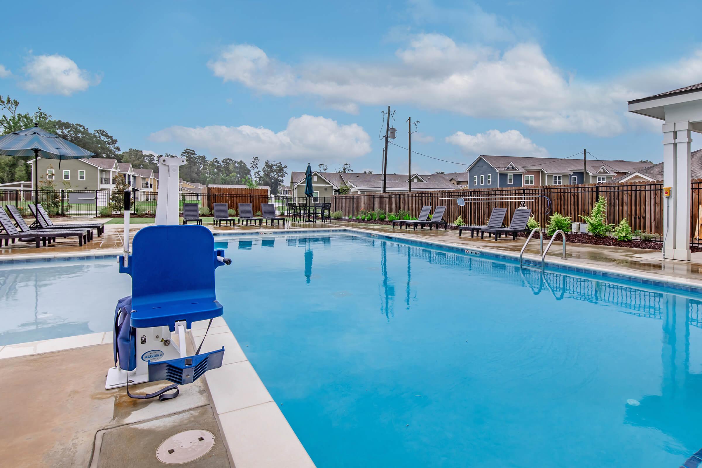 A clear swimming pool surrounded by lounge chairs and umbrellas on a sunny day. The pool area features a ramp for accessibility, with residential buildings visible in the background. The sky is bright blue with a few clouds, creating a serene atmosphere.
