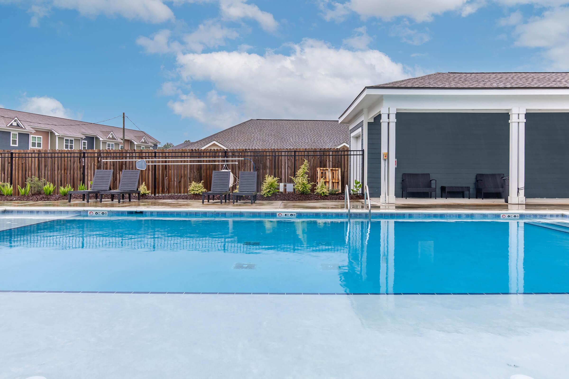 A clear blue swimming pool with lounge chairs on the deck, surrounded by a wooden fence. There is a building in the background with a modern design, and the sky is bright with fluffy white clouds. Lush greenery is visible along the pool area, creating a relaxing atmosphere.