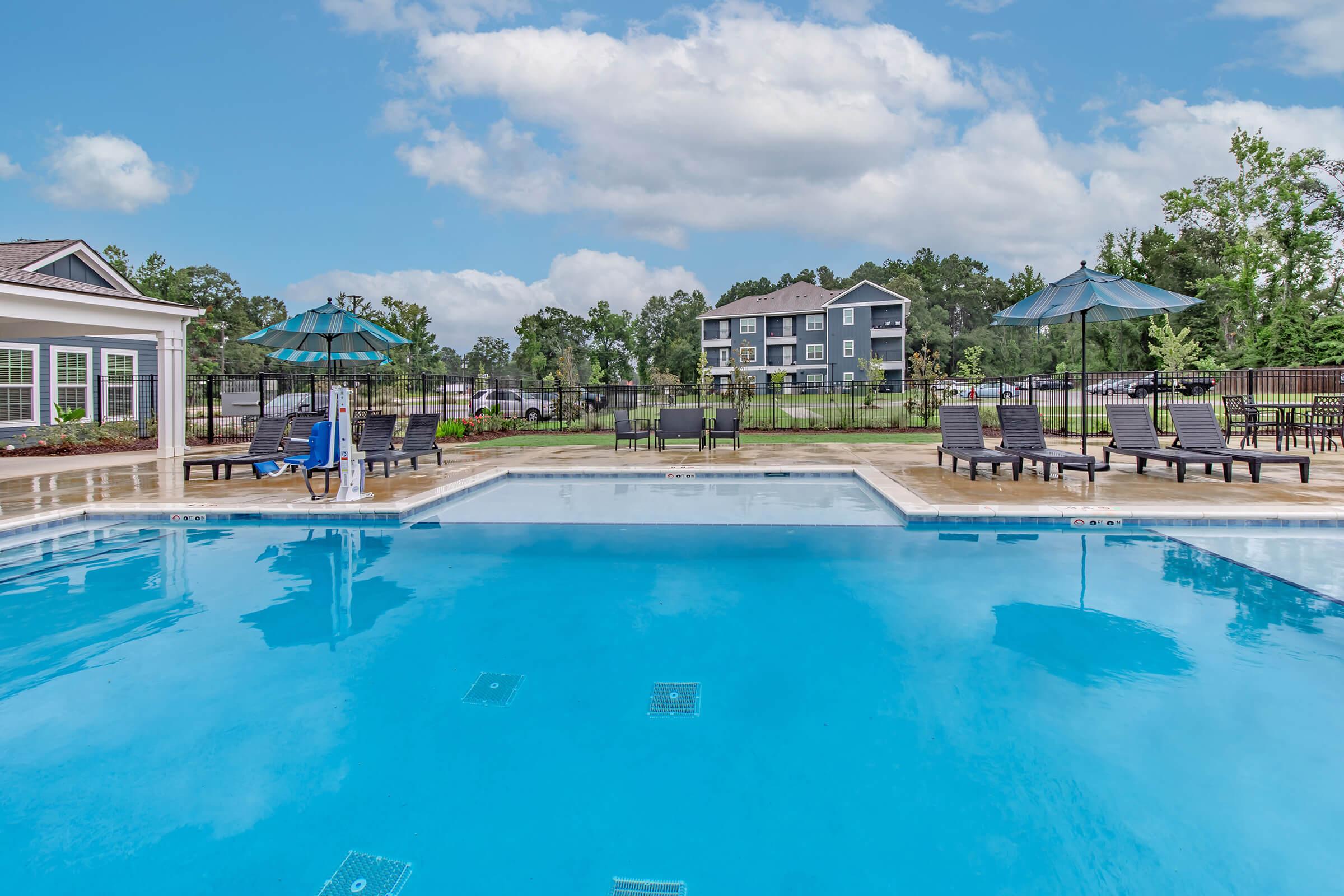 A clear blue swimming pool with lounge chairs and umbrellas around it. In the background, there are green trees and a multi-story apartment building. The sky is partly cloudy, creating a bright and inviting atmosphere.