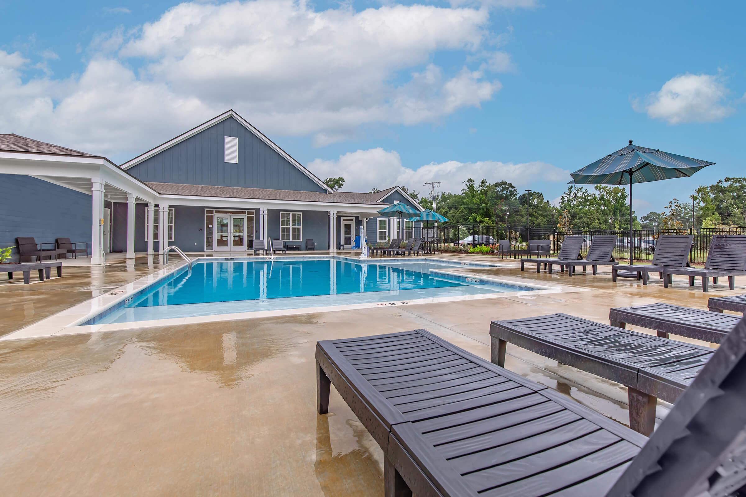 A clear blue swimming pool surrounded by wooden lounge chairs, with a large umbrella providing shade. In the background, there's a modern building with a gray exterior and white trim. The sky is partly cloudy, and the area appears well-maintained and inviting.
