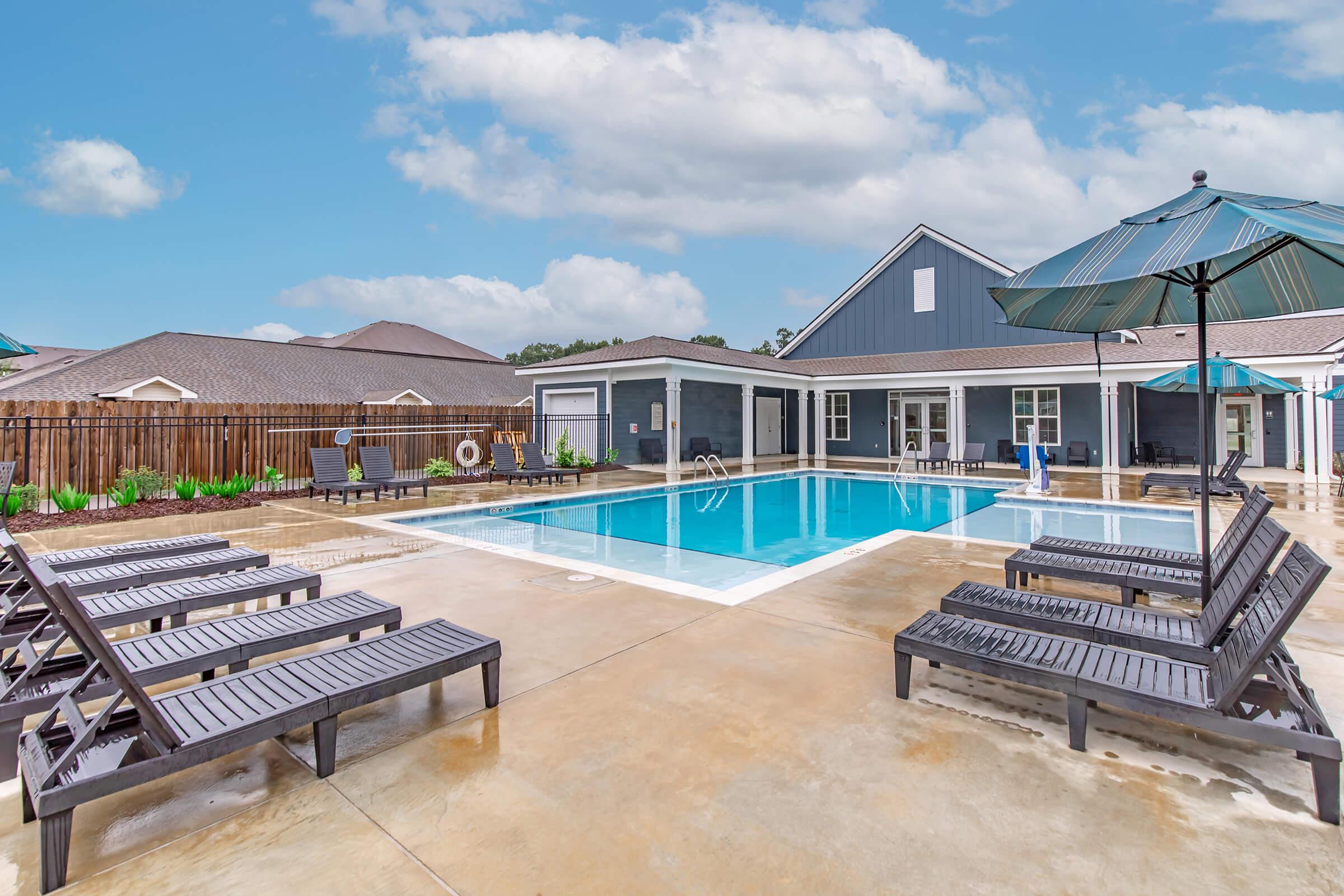A spacious outdoor pool area featuring a rectangular swimming pool surrounded by lounge chairs and shaded by umbrellas. The deck is wet, suggesting recent rain, and there are modern buildings in the background, creating a relaxed atmosphere. Cloudy blue skies add to the serene setting.