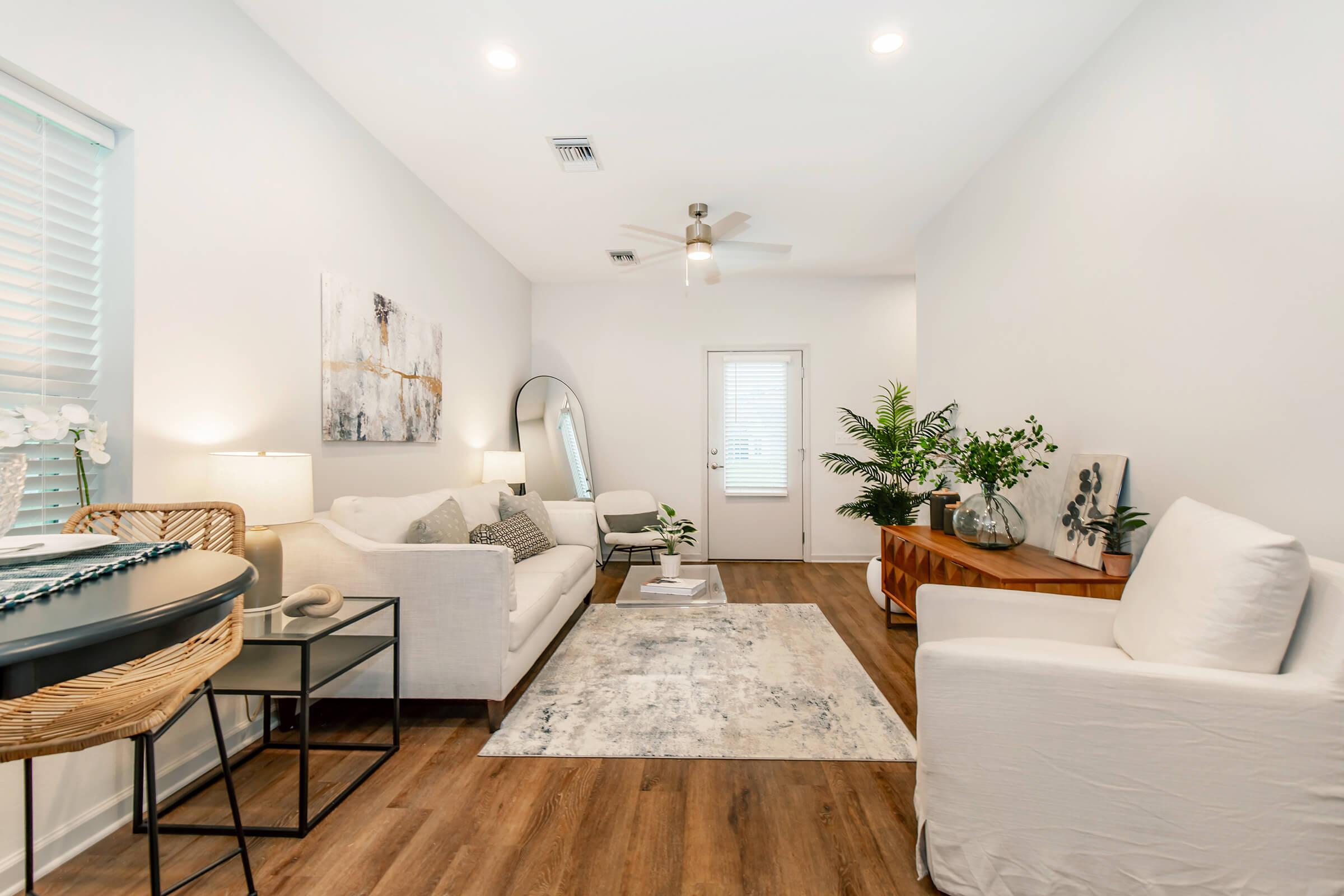 A cozy, modern living room featuring a white sofa, a light-colored armchair, and a round dining table. A large mirror and decorative plants enhance the space, complemented by a neutral area rug on wooden flooring. Natural light streams in through a window, creating a warm and inviting atmosphere.