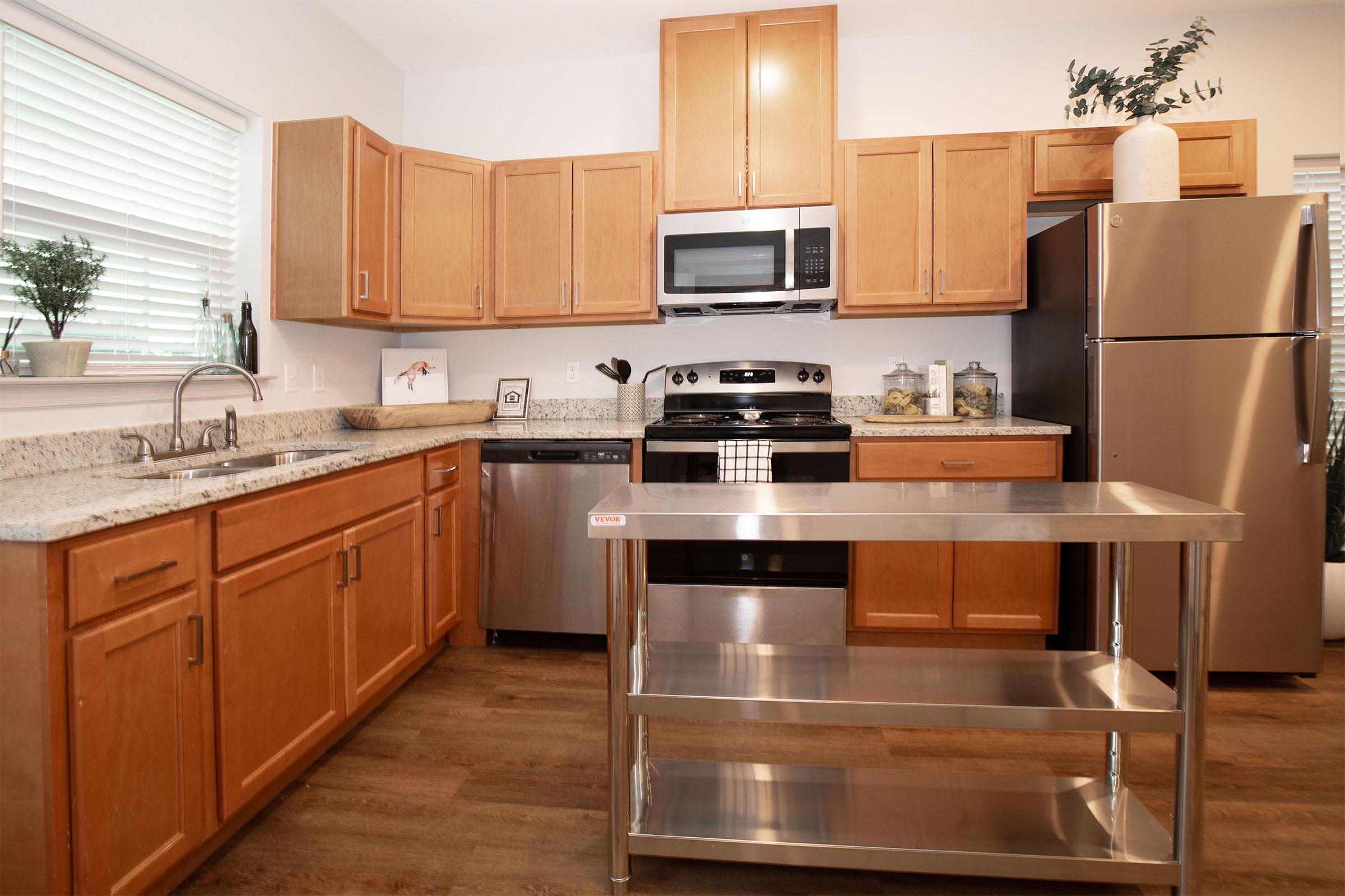A modern kitchen featuring wooden cabinets, stainless steel appliances, and a central metal kitchen island. The countertop is made of light granite, and there are some decorative plants in pots. Large windows provide natural light, enhancing the inviting and clean atmosphere.