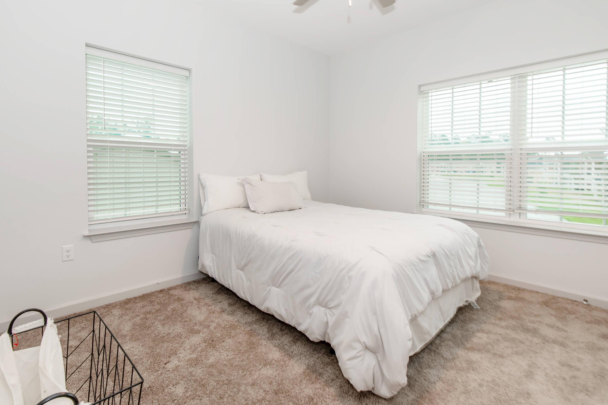 A simple, clean bedroom featuring a white bed with a plush comforter, two windows with blinds allowing diffused light, and a neutral carpet. A black wire basket is positioned on the floor, adding a touch of minimalist decor to the space. The walls display a light, calming color.