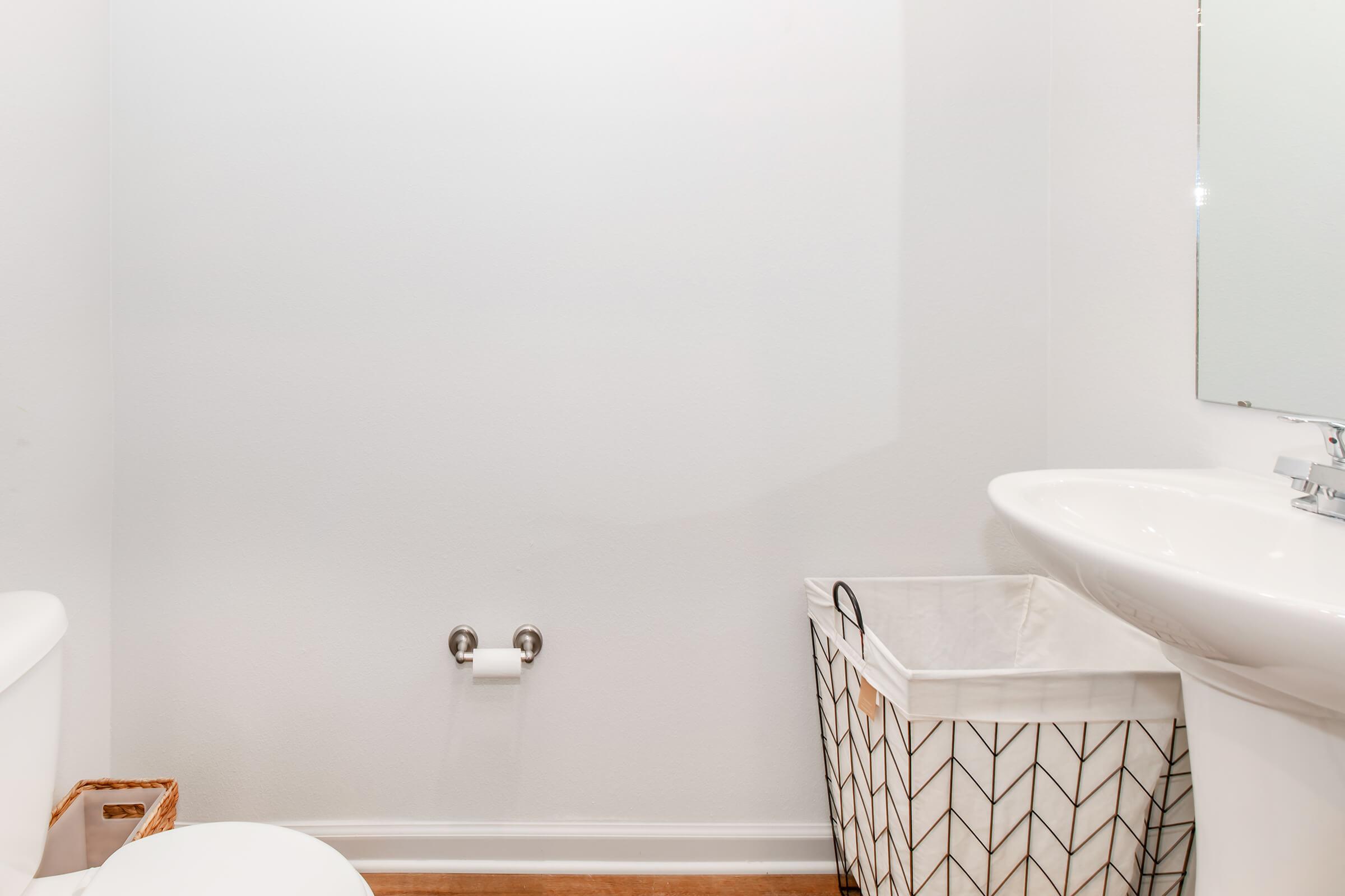 A clean bathroom interior featuring a white wall, a toilet, a sink with a modern faucet, and a mirror. To the right, there is a patterned laundry basket. The floor is wooden, and the overall space has a bright and minimalist design. Stability and simplicity characterize the room.