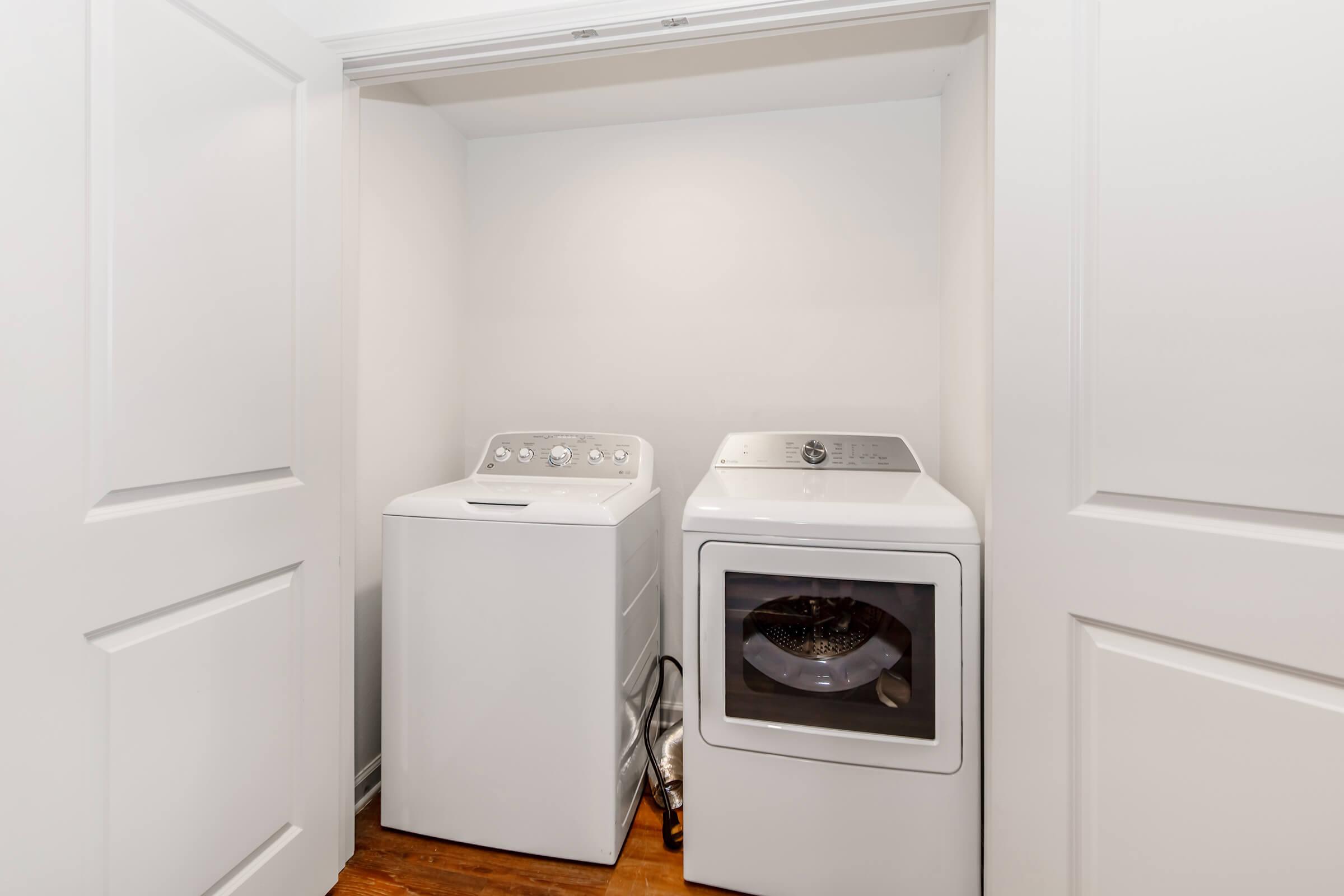 A compact laundry area featuring a white washing machine and a white dryer side by side in a small closet with white doors. The floor is wooden, and the overall space is clean and organized, ideal for home laundry needs.