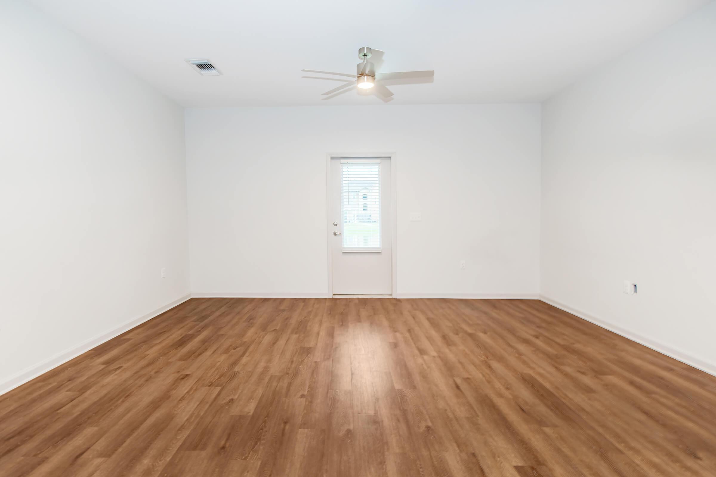 A spacious, empty room with light-colored walls and laminate flooring. A ceiling fan is visible, and there is a door with a window leading outside. The room is well-lit by natural light coming through the door.