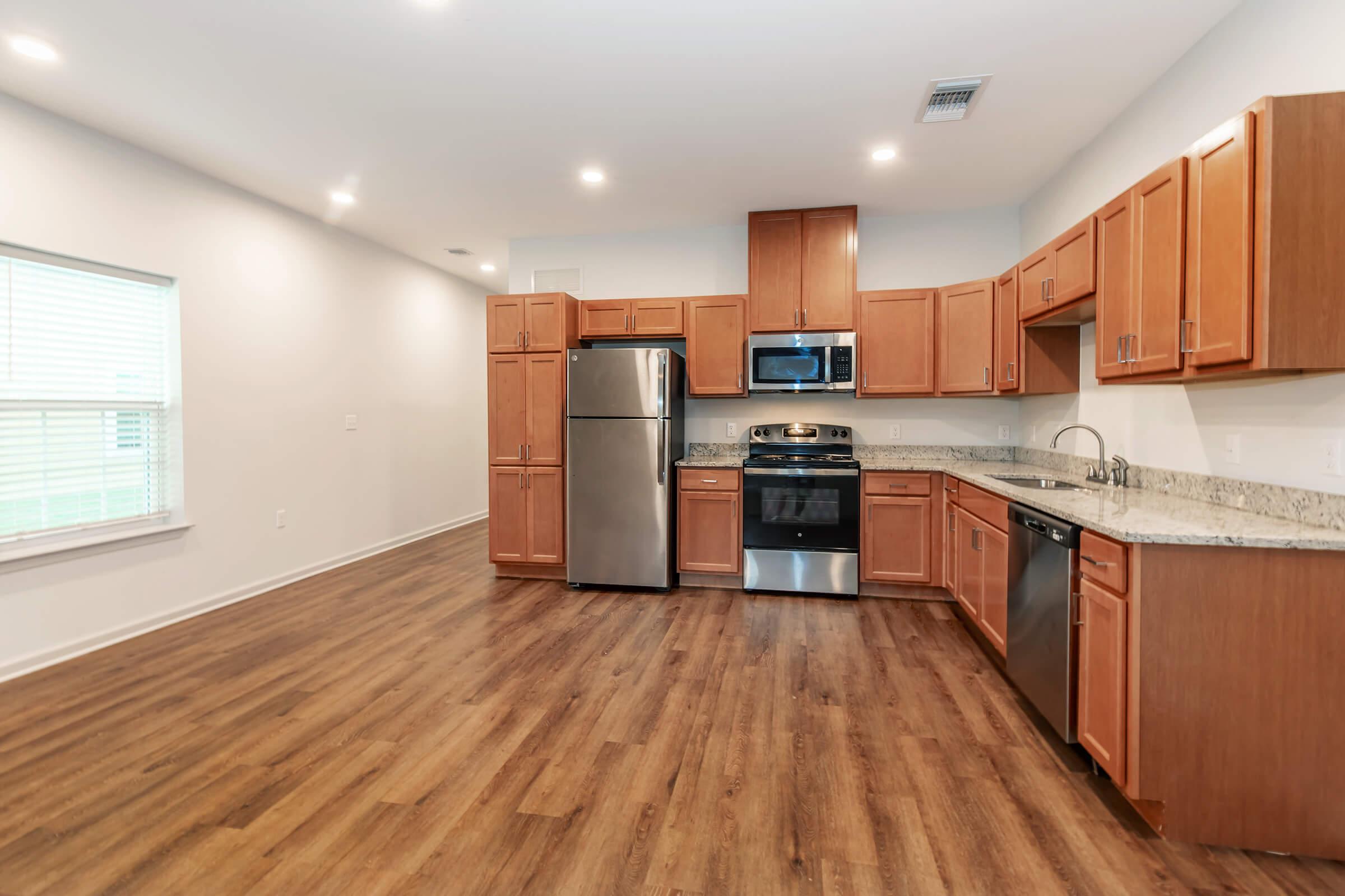 A modern kitchen featuring stainless steel appliances, including a refrigerator, stove, and dishwasher. The cabinetry is made of light wood, and the countertop is granite. The space has a wood-style floor and large windows that allow natural light to brighten the room.