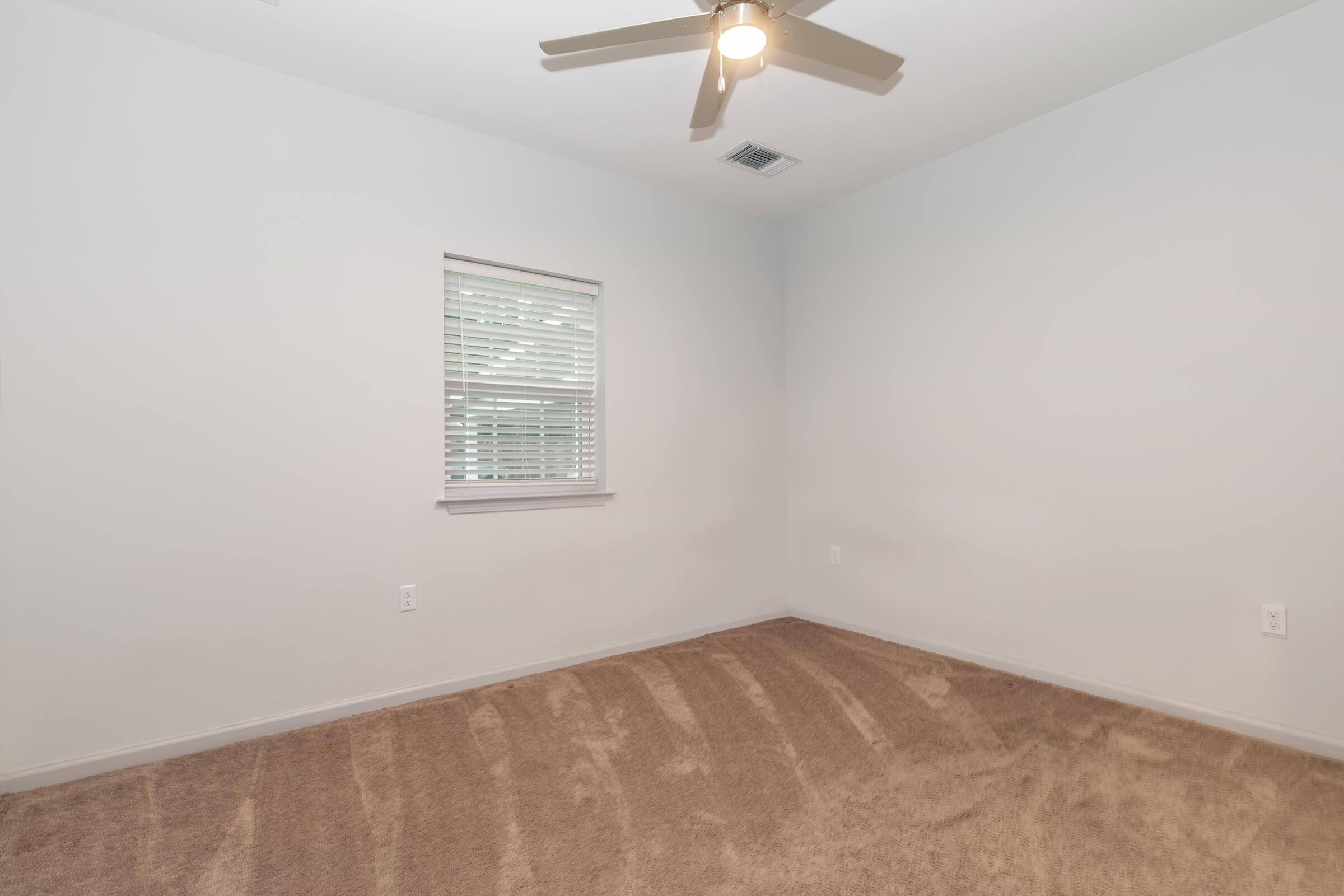 Empty room with light gray walls and a ceiling fan. A small window with blinds is visible, allowing natural light to enter. The floor is covered with beige carpet, and there are no furnishings or decorations. The overall appearance is clean and minimalistic.