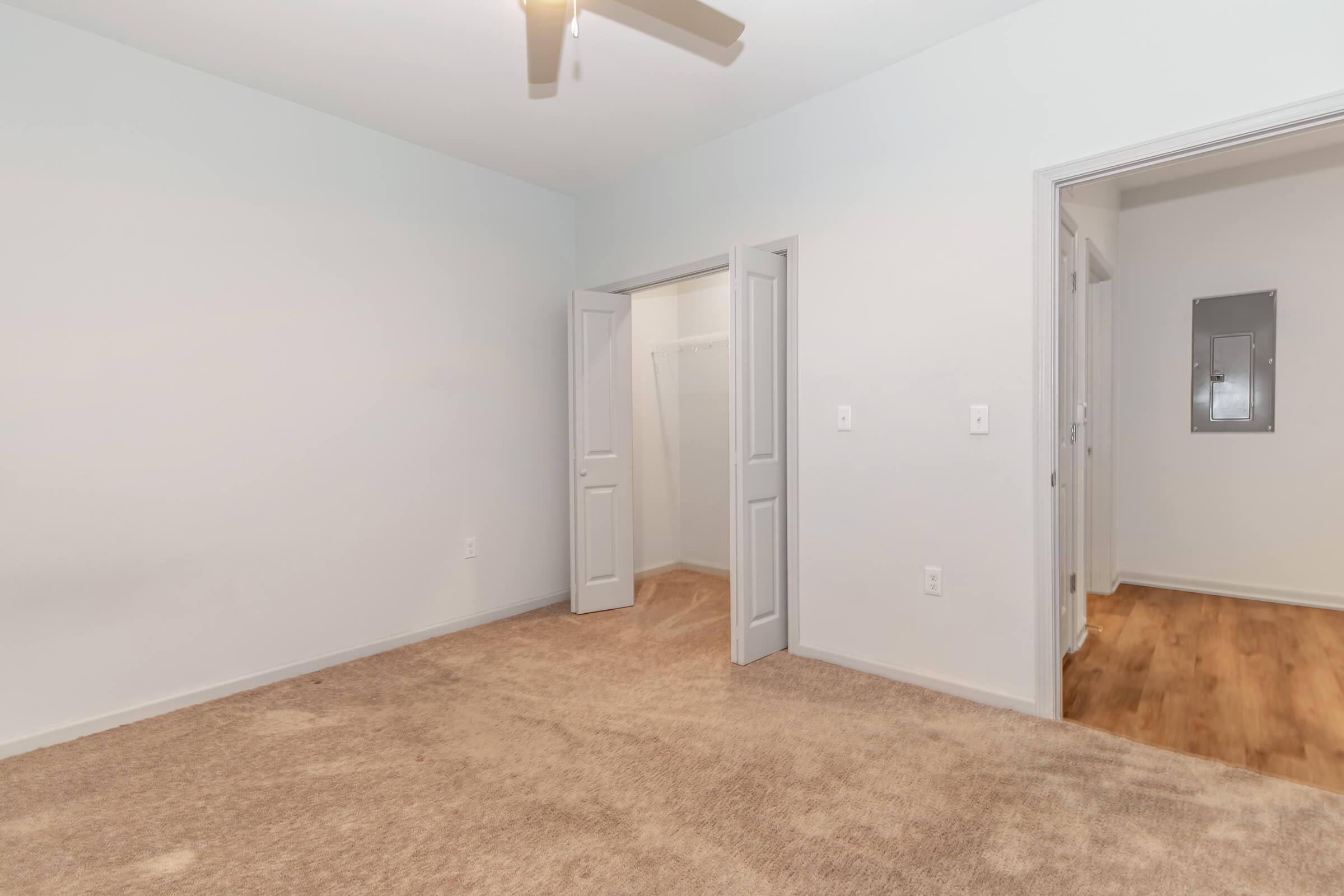 A vacant room with beige carpet flooring, light gray walls, and a ceiling fan. There's an open doorway leading to a small closet on the left and another door on the right, likely leading to a hallway. Natural light coming through the doorway gives the space a bright appearance.