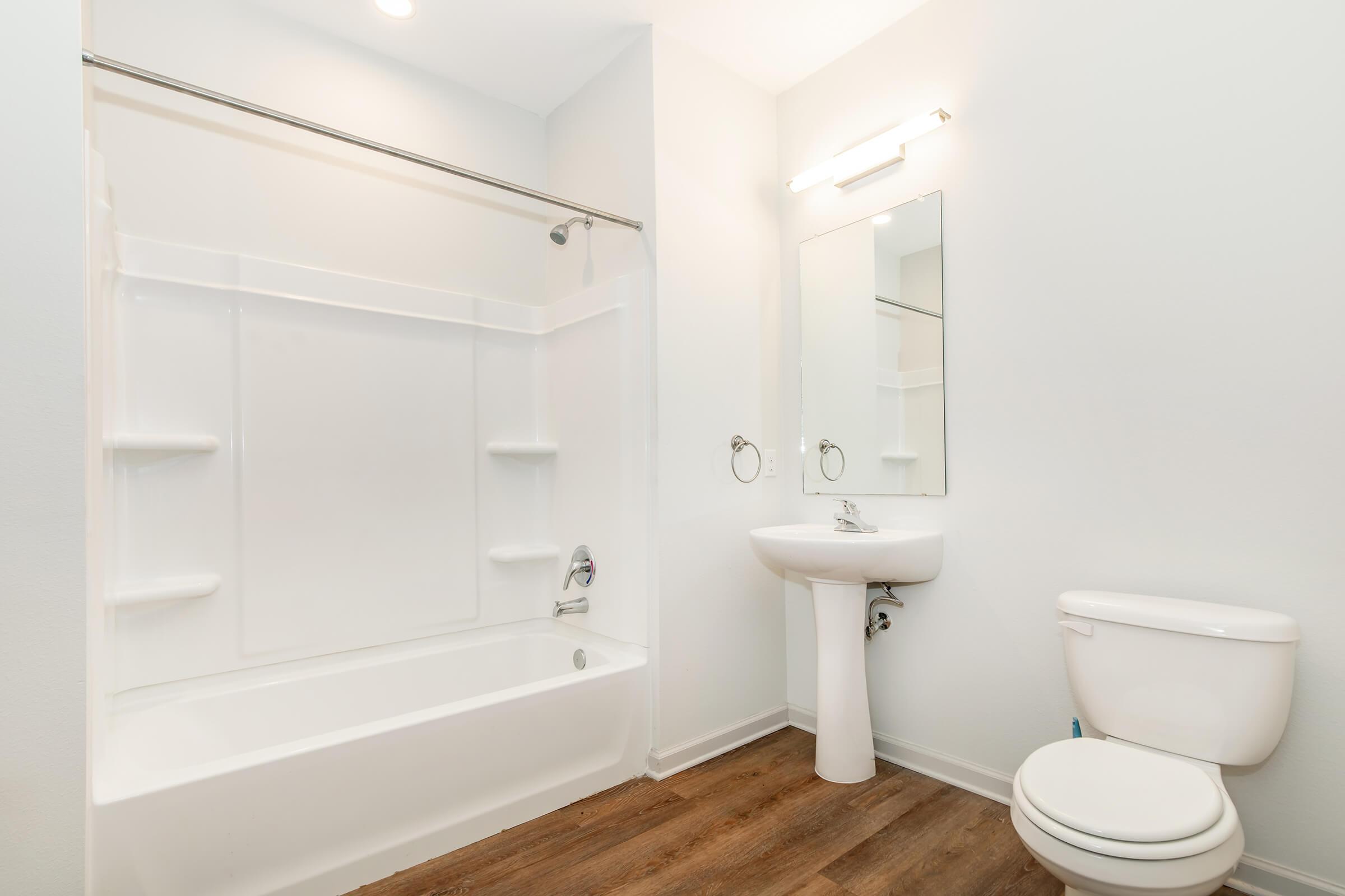 A clean, modern bathroom featuring a white bathtub with a shower, a pedestal sink with a mirror above, and a toilet. The walls are painted white, and the floor has a warm wood-like finish, creating a bright and spacious atmosphere.