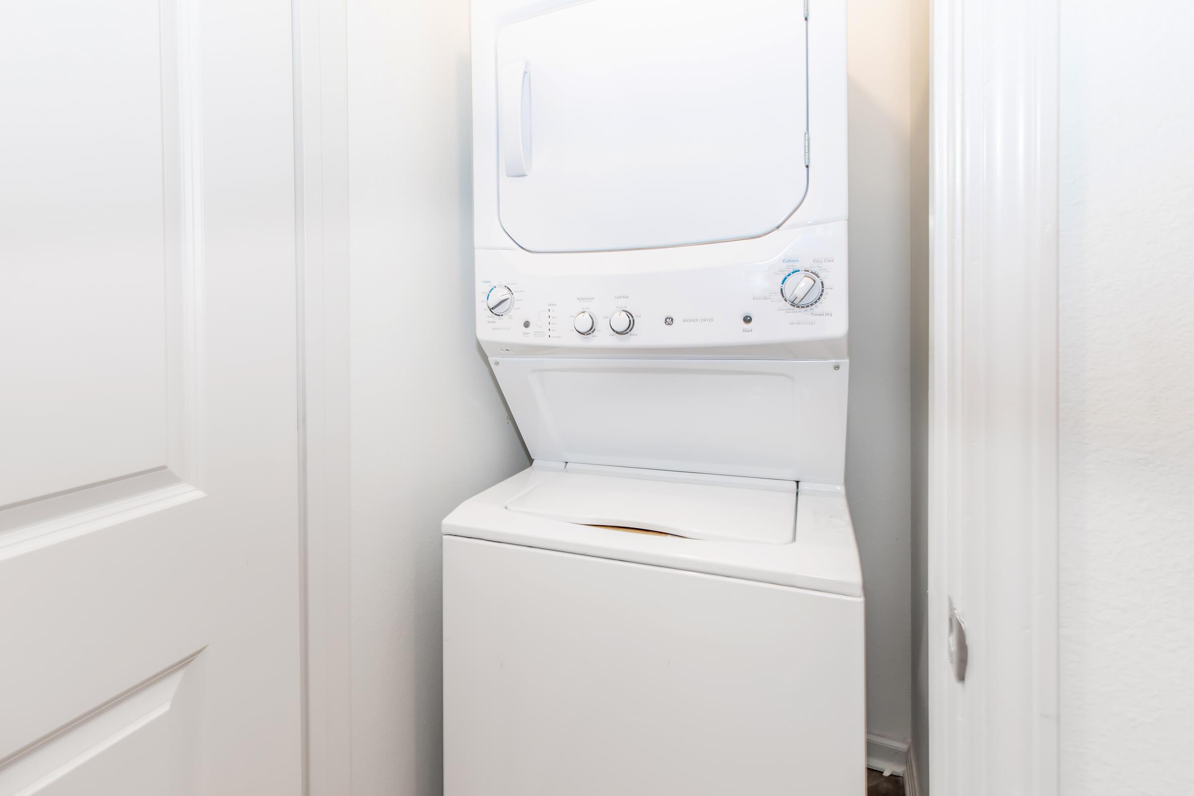 A stacked washer and dryer unit located in a narrow space, with a white exterior. The dryer is on top, featuring dials and controls on the front panel, while the washing machine is beneath it. The surrounding area includes plain white walls and a closed door nearby.