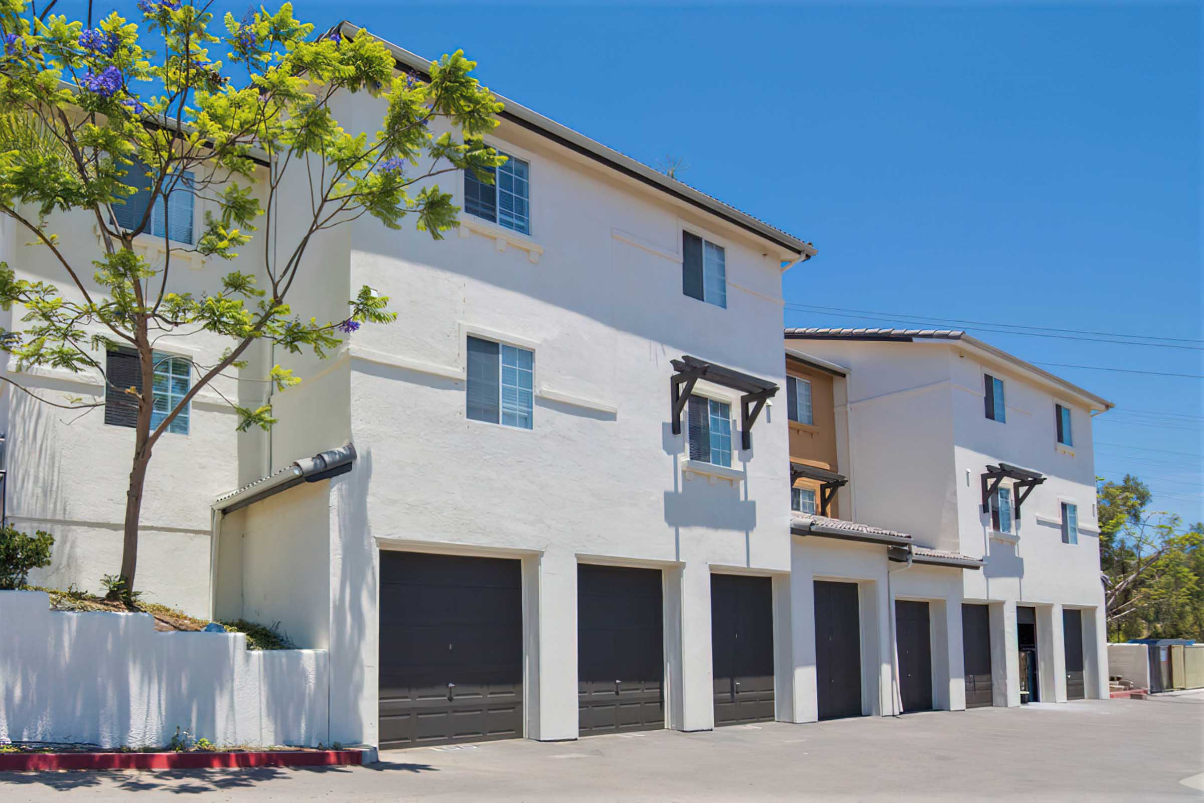 A row of three multi-story residential buildings with a white exterior, featuring balconies, large windows, and garage doors at the ground level.