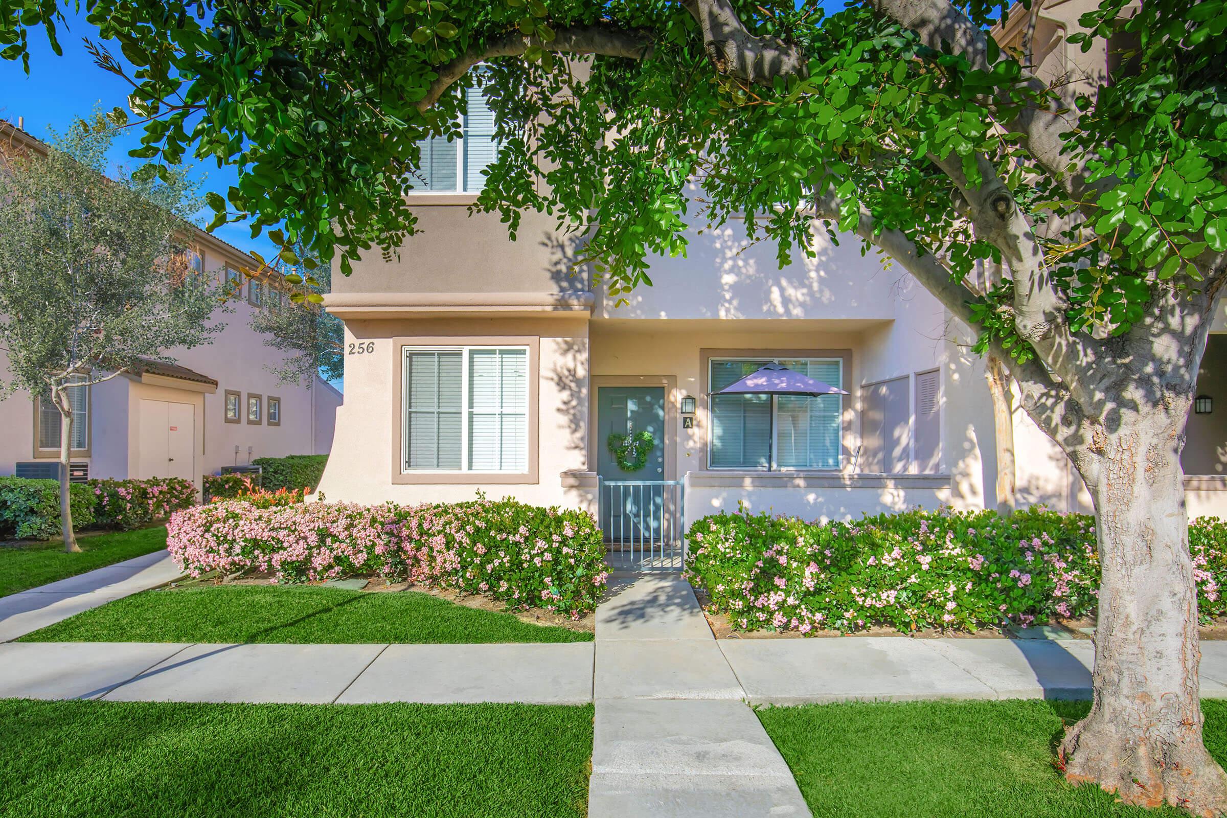 Front view of a light-colored house with a well-maintained garden. It features a small porch with a green door, surrounded by blooming flowers and manicured grass. A large tree is in the foreground, providing shade to the walkway leading to the entrance. Sunny day with clear blue skies.