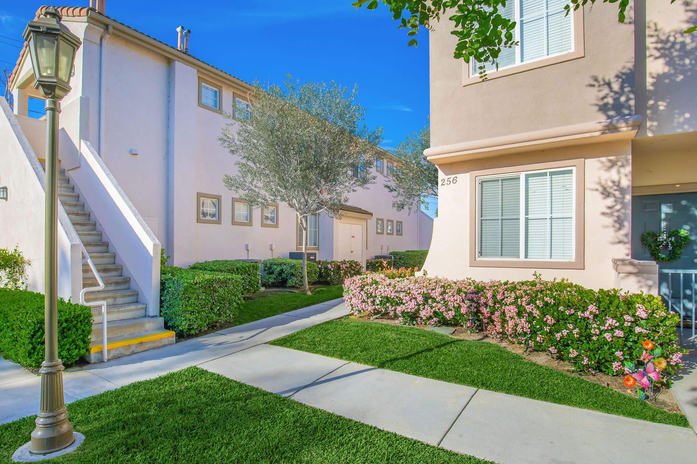 A well-maintained residential area featuring two pastel-colored buildings with stairs leading up to one side. The path is surrounded by lush green grass and vibrant flower beds, creating a pleasant and inviting atmosphere. A street lamp stands near the walkway, adding to the charm of the scene.