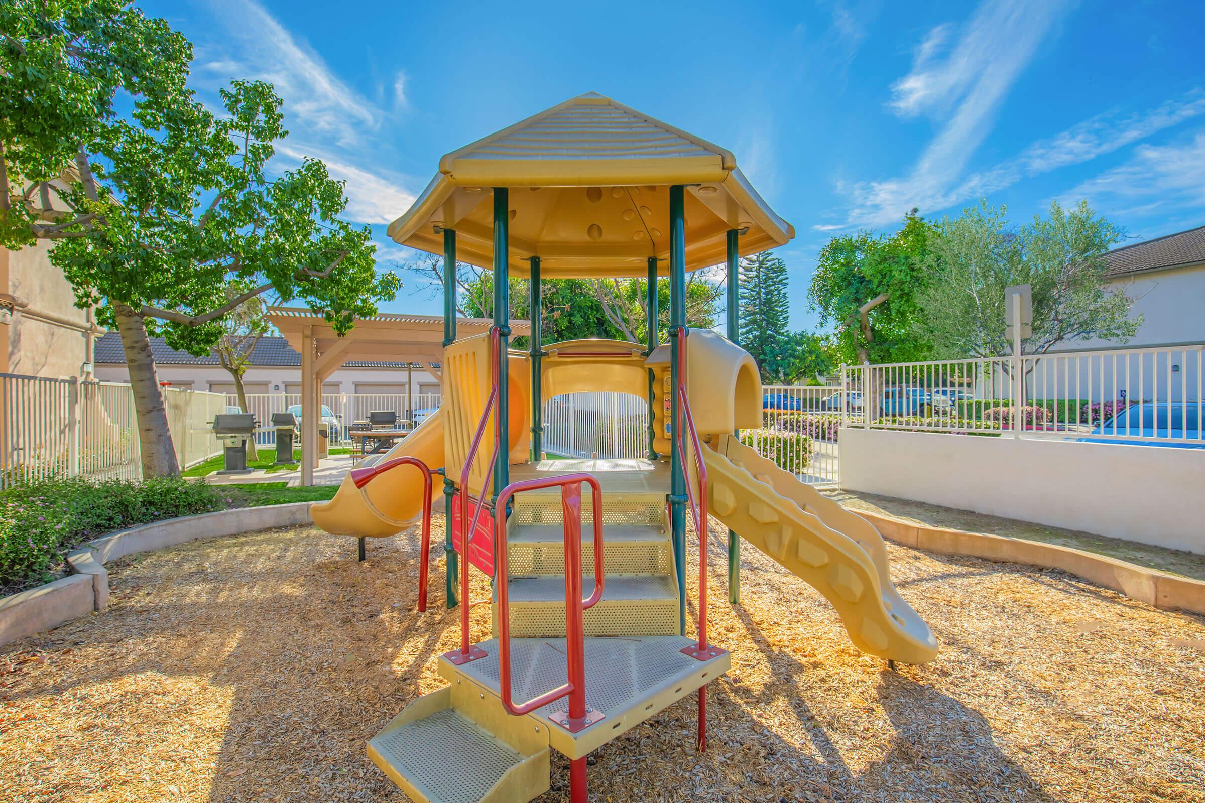A colorful playground structure featuring a slides and stairs, surrounded by a sandy area. Lush green trees and a clear blue sky with a few clouds can be seen in the background, along with a white fence enclosing the play area.