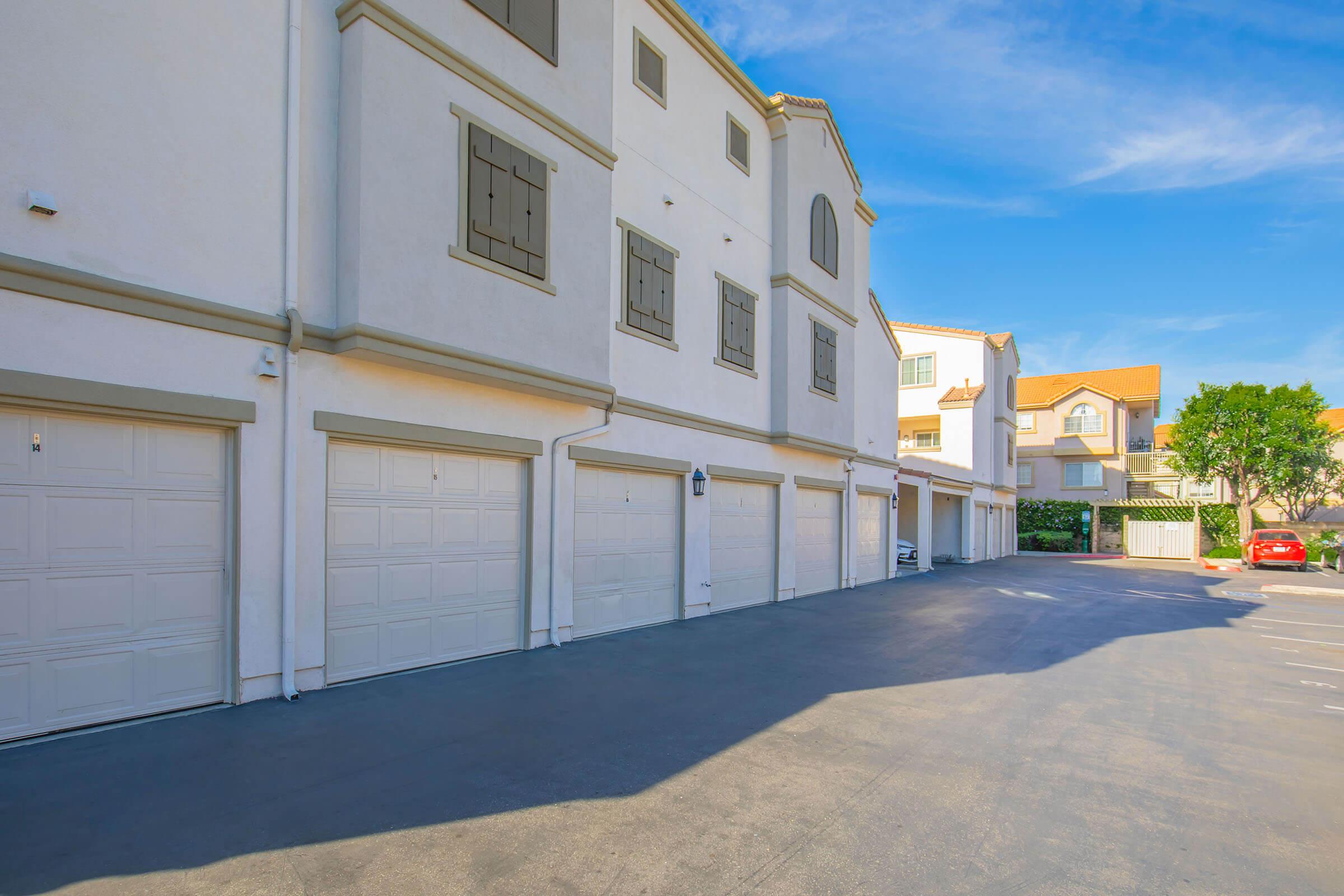 A row of garage doors attached to a residential building. The white stucco exterior features decorative elements and windows, under a clear blue sky. The area is paved with asphalt, and additional buildings and trees are visible in the background.